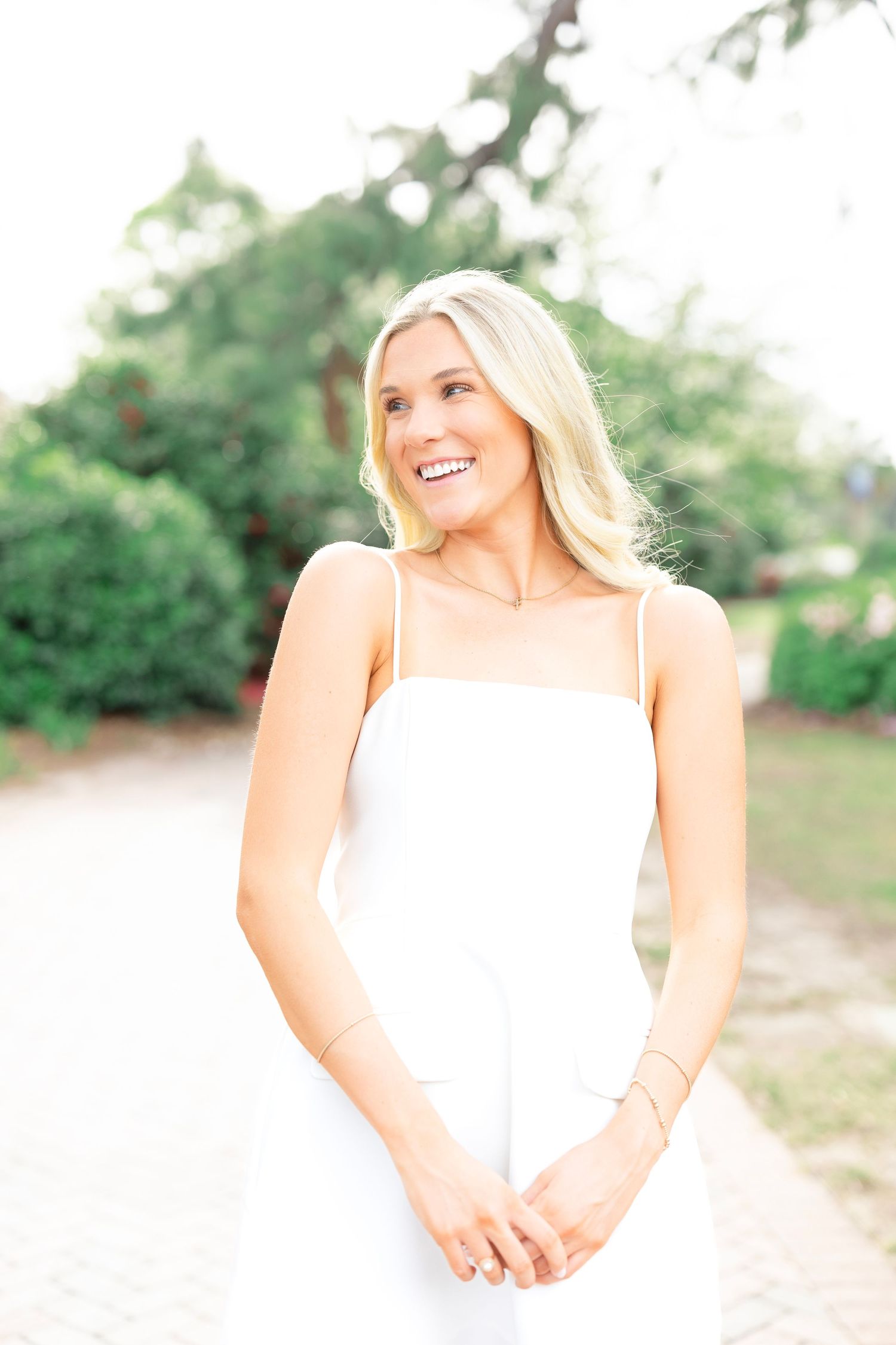 A woman in a white sleeveless dress poses outdoors in natural lighting.