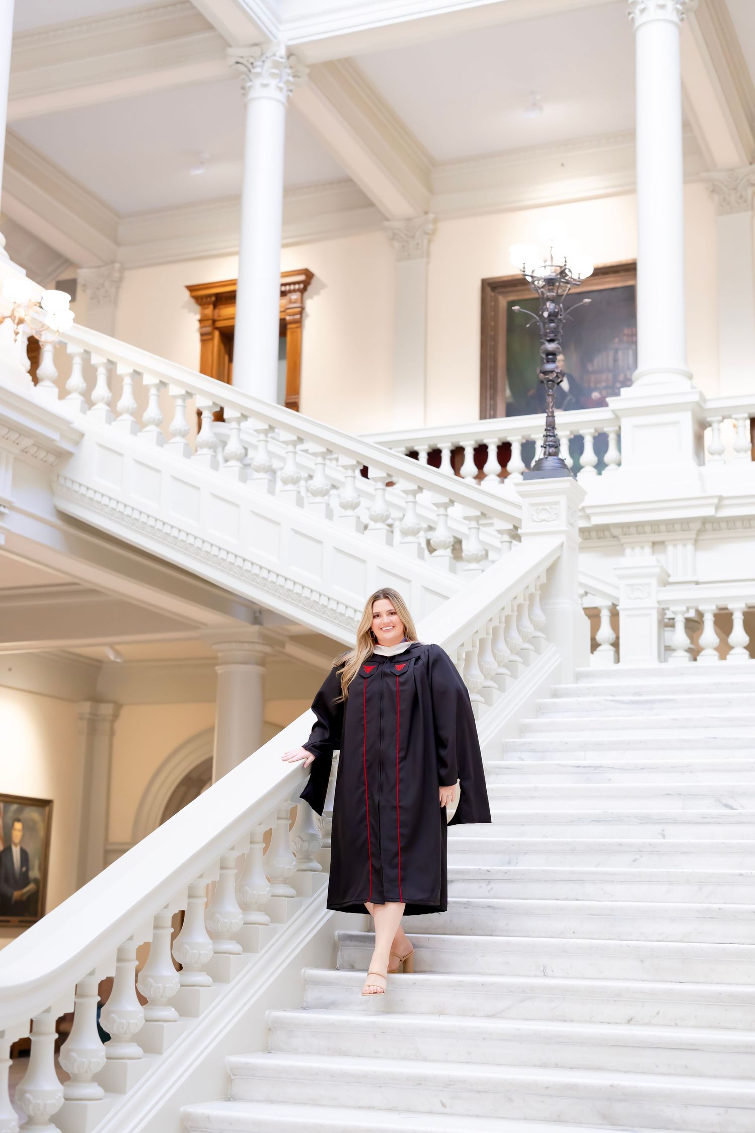 Graduate in academic regalia poses on grand white marble staircase in elegant building interior.