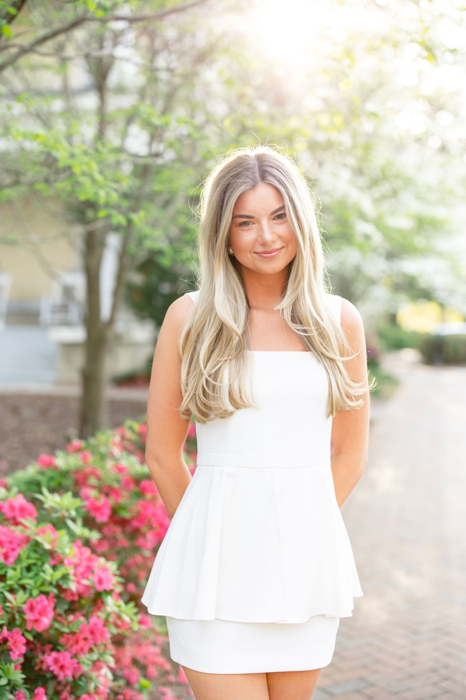 A woman in a white sundress stands on a garden path surrounded by pink azalea bushes.