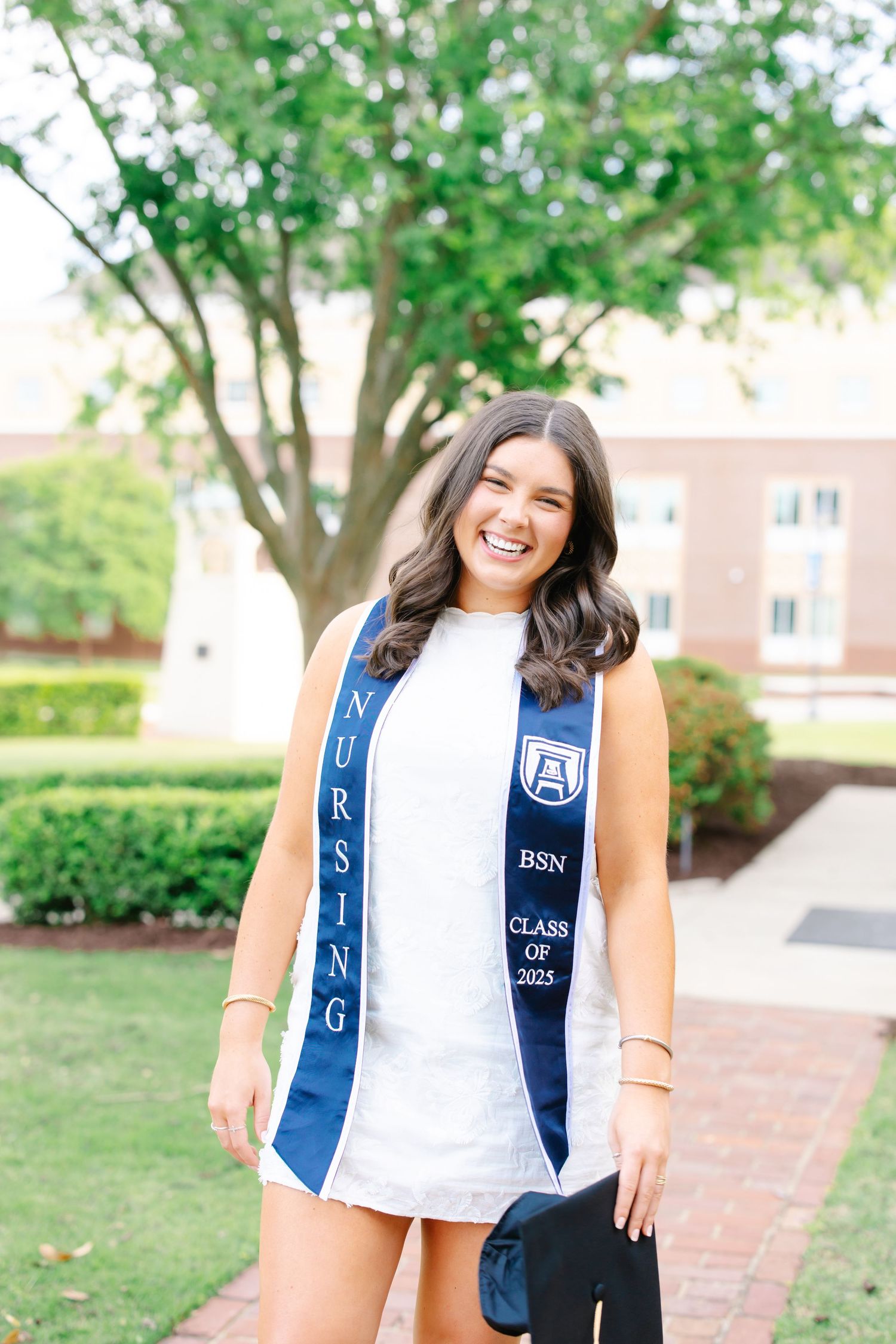 Nursing graduate in white dress and blue stole smiles while holding graduation cap outdoors.