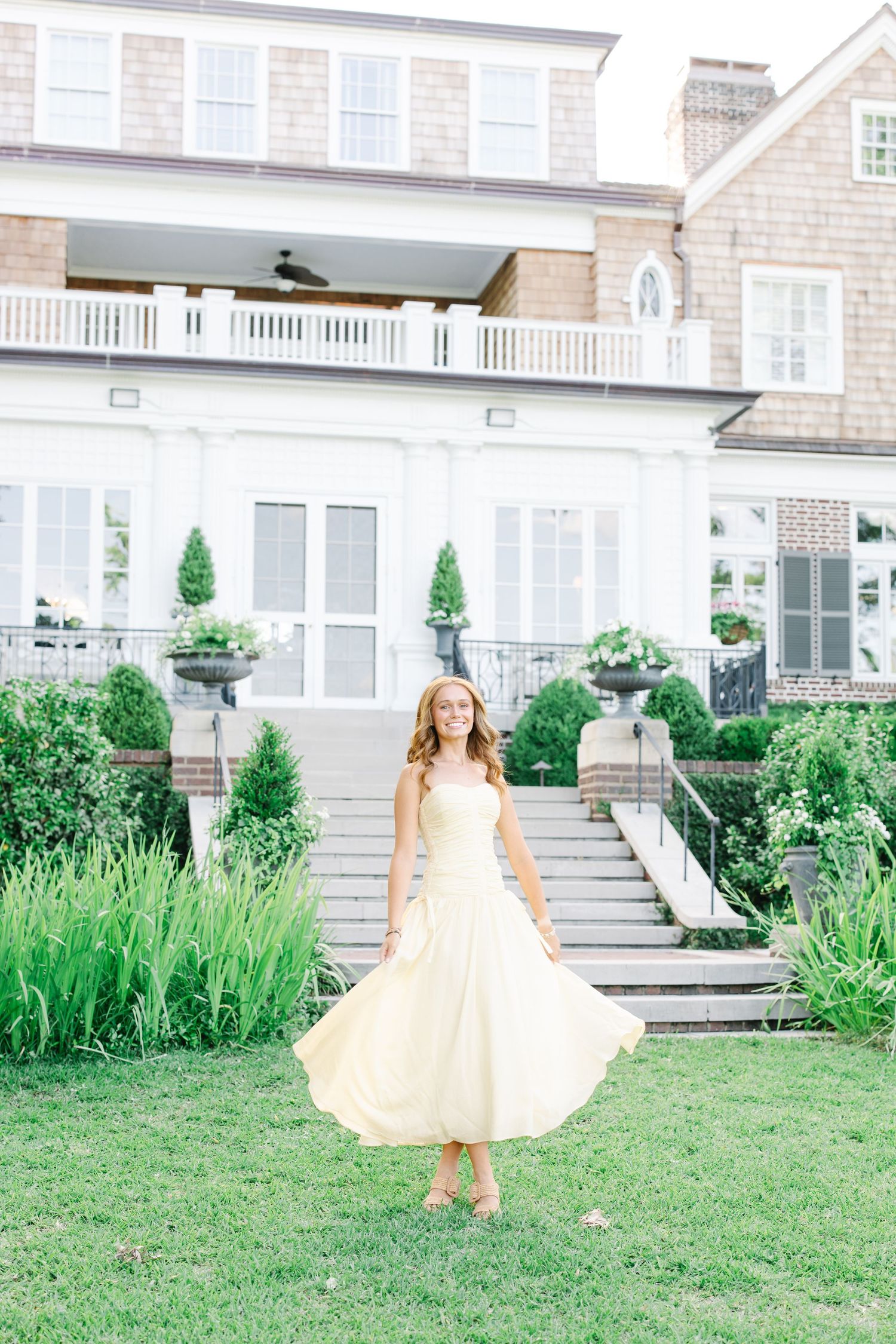 A woman in a yellow sundress twirls on a manicured lawn in front of a stately home with white columns.