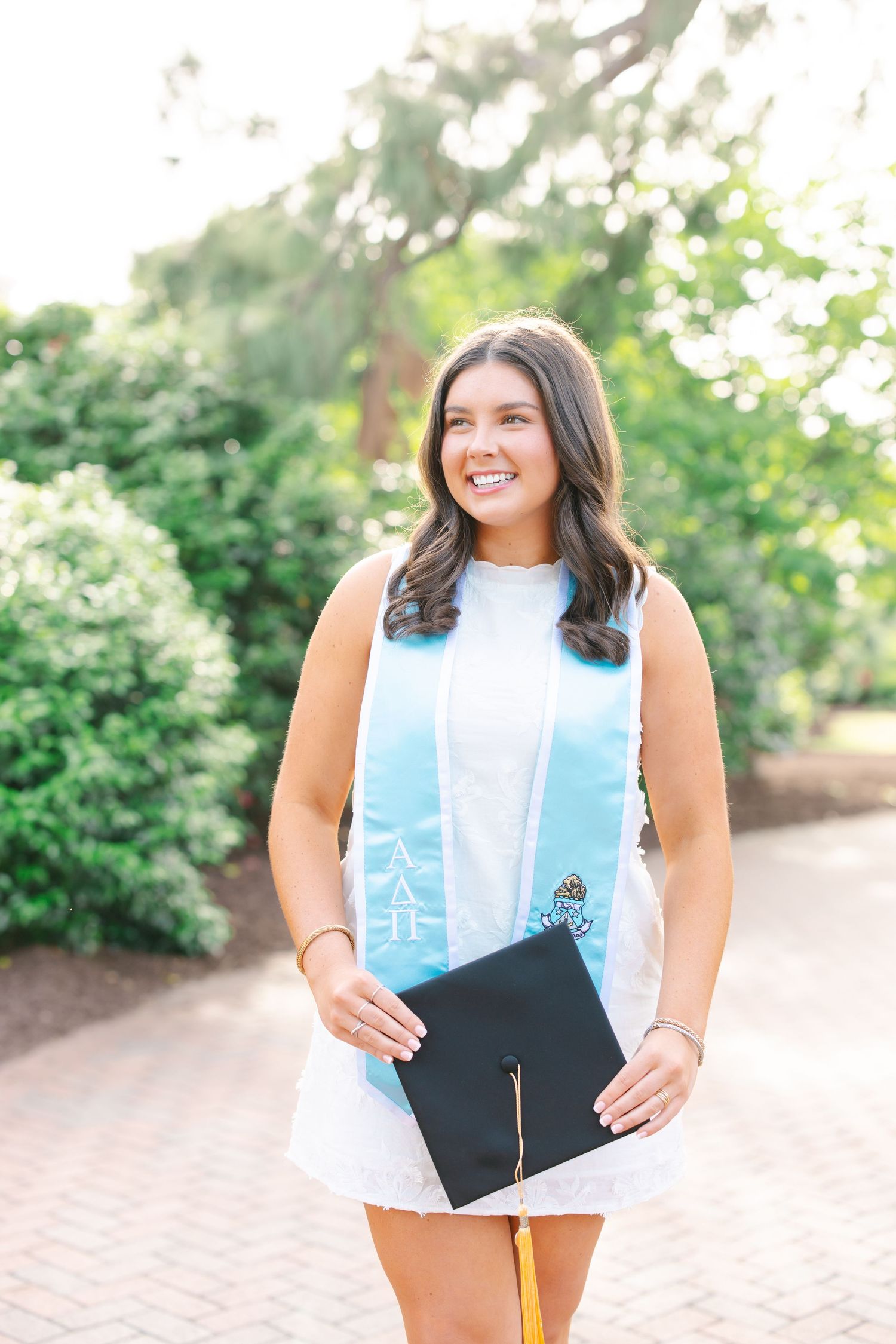 Graduate in white dress and light blue graduation stole holding diploma on campus.