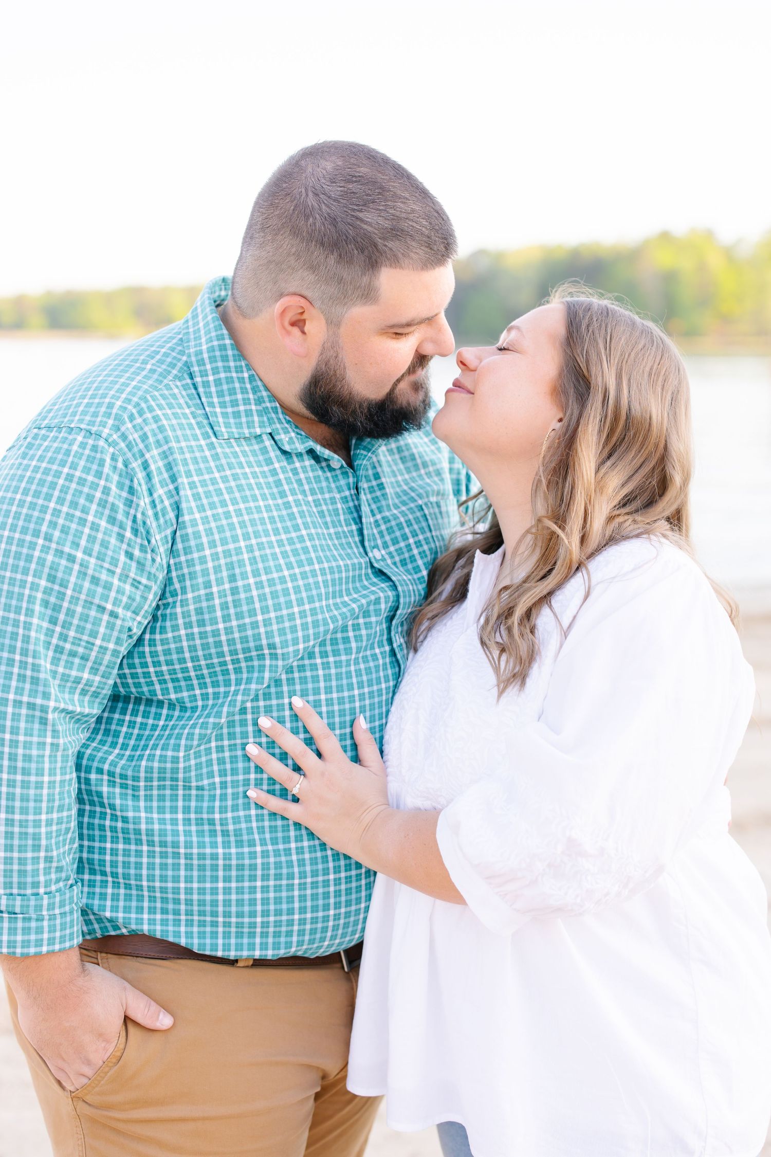 A couple shares an intimate embrace while showing off an engagement ring by the water.