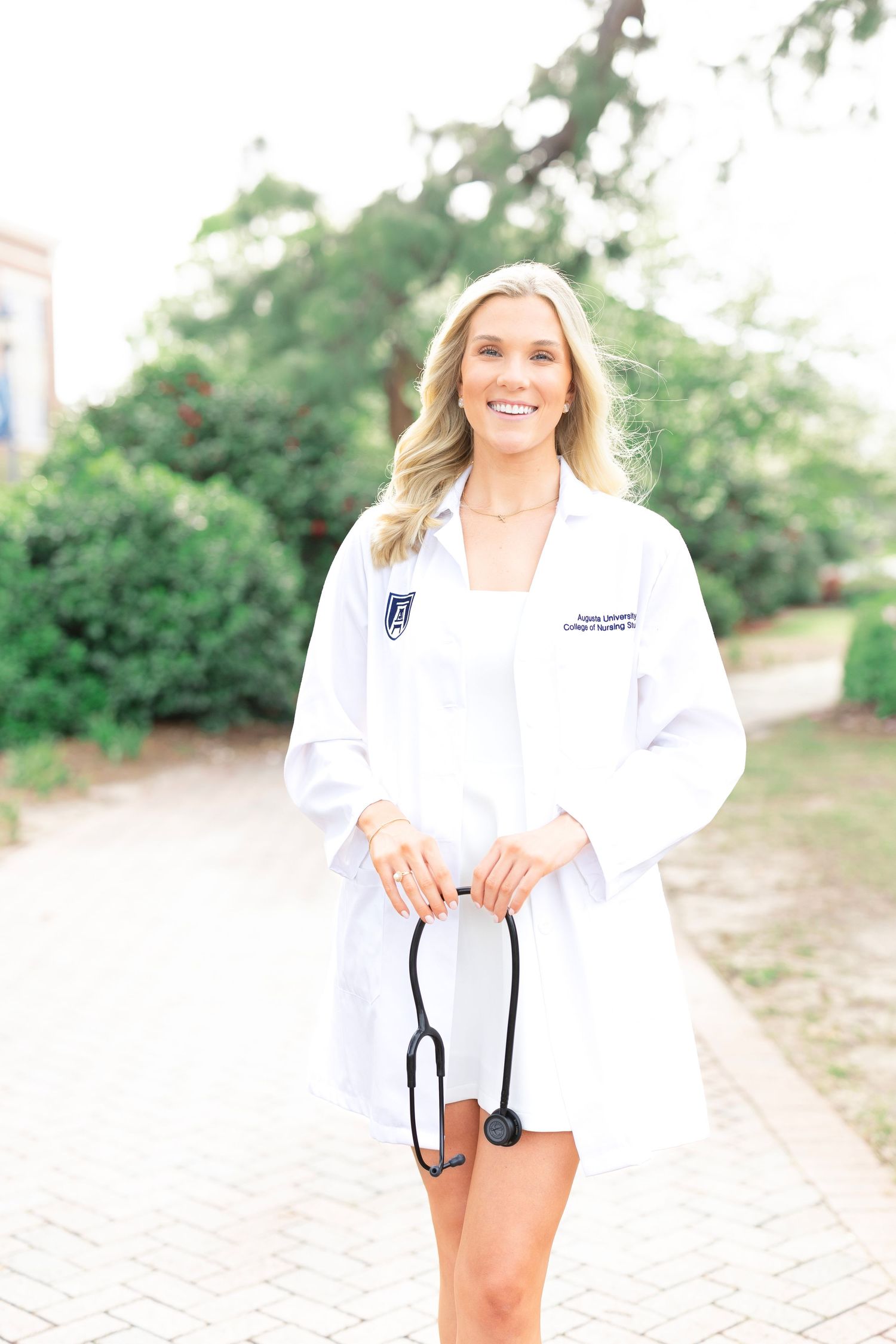A medical professional in a white coat holds a stethoscope while standing on a brick pathway.