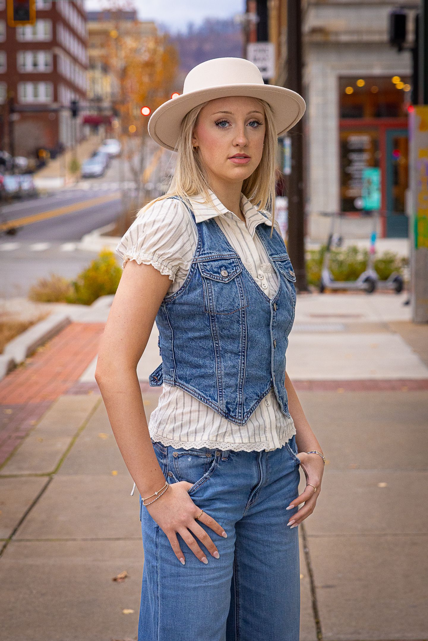 Blonde woman in denim overalls vest, white blouse, and hat poses on urban sidewalk in a stylish casual outfit.