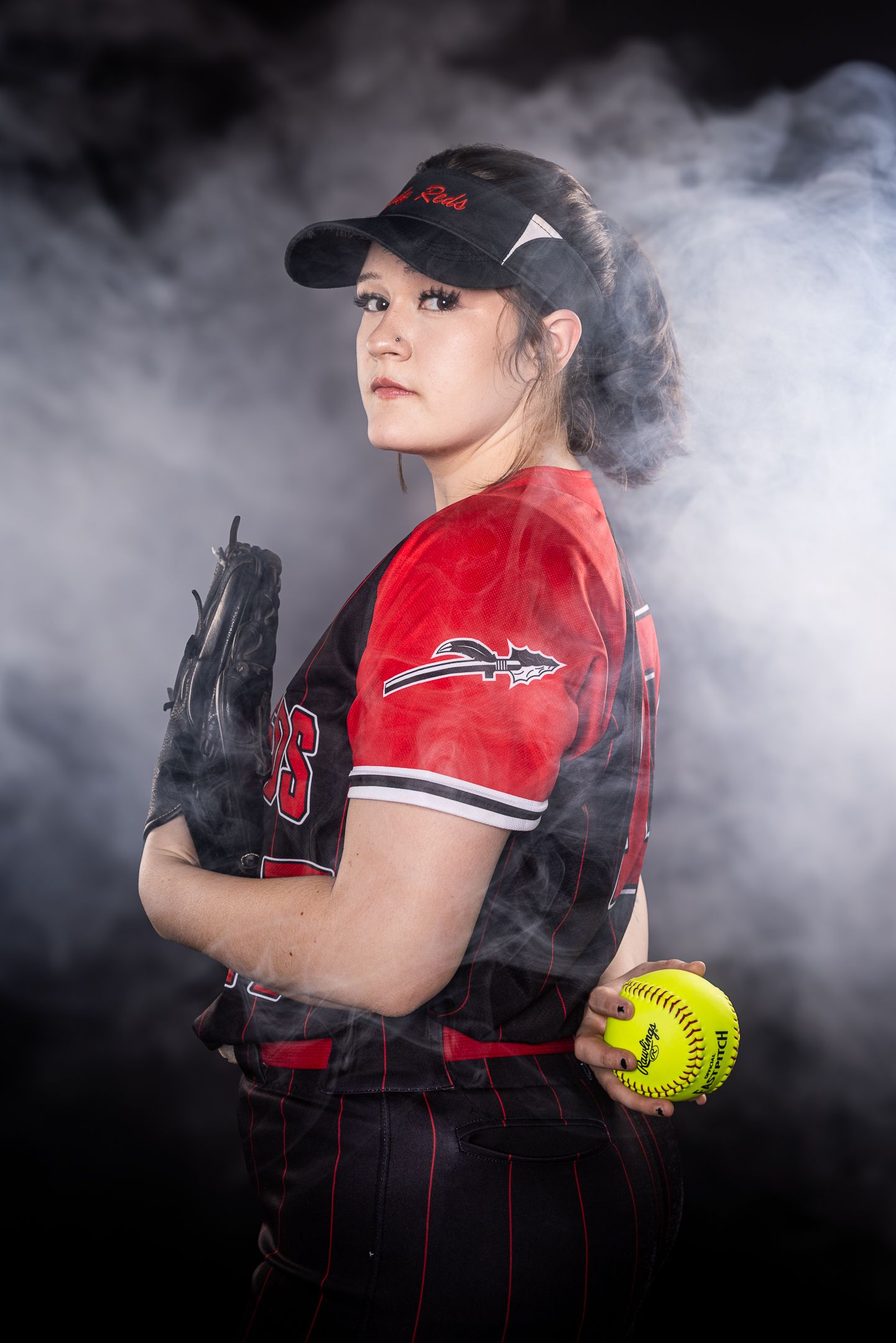 Softball player in red uniform poses with ball and glove amid atmospheric smoke effects.
