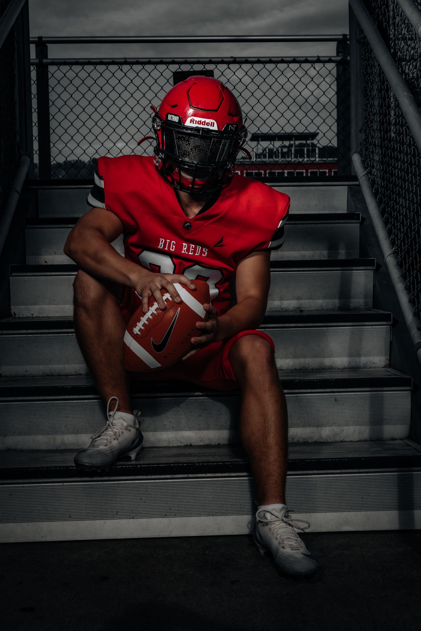 Football player in red uniform sits on stadium steps in dramatic lighting.