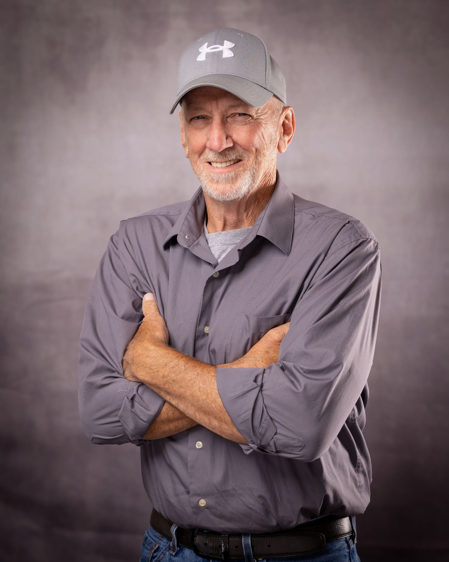 Smiling older man with crossed arms wearing a gray Under Armour cap and button-up shirt in a studio portrait.