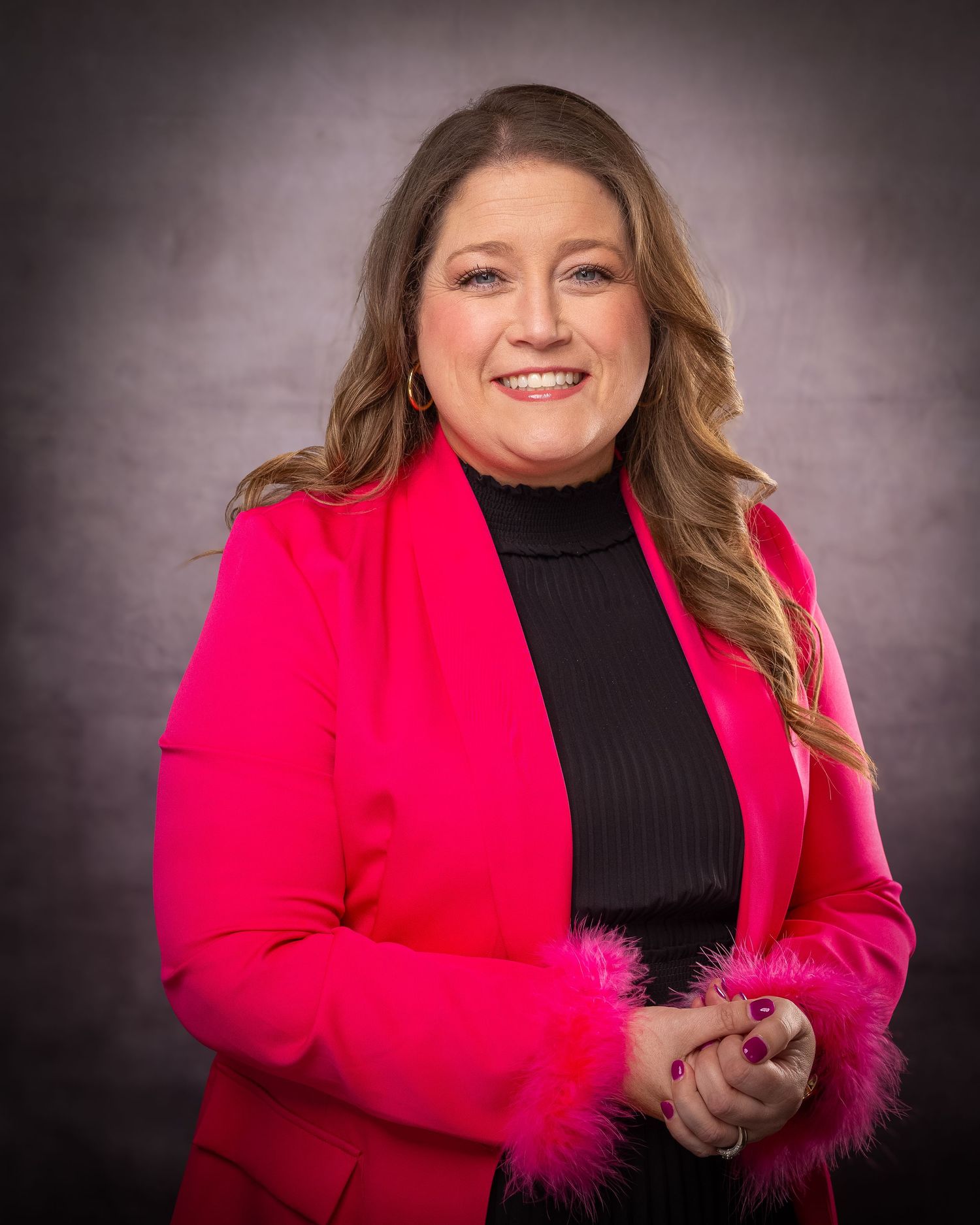 Woman in hot pink blazer with feather cuffs and black turtleneck smiling confidently against gray studio background.