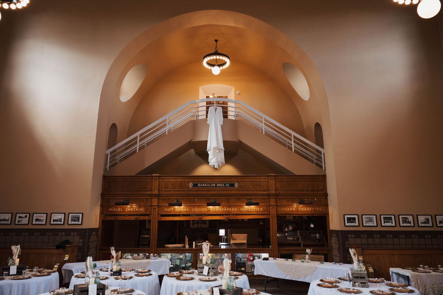 A white wedding dress hangs elegantly in an arched cathedral ceiling venue with round tables set for a reception.