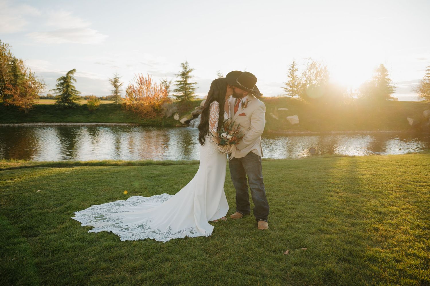 A couple in wedding attire embraces during sunset near a lake with golden light filtering through trees.