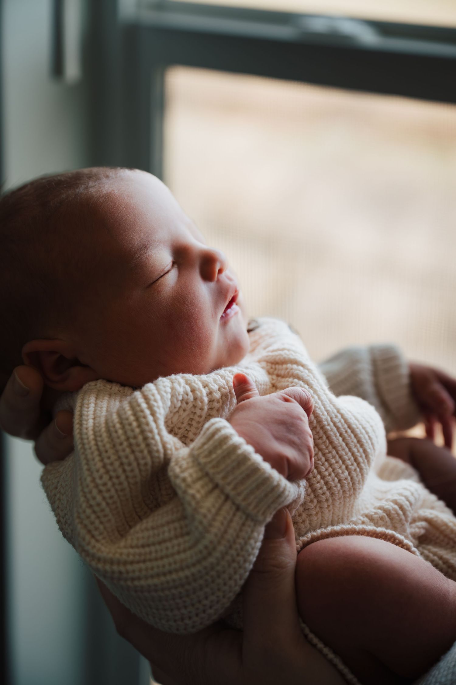 A peaceful sleeping baby dressed in a cozy cream knit sweater rests near a window in natural light.
