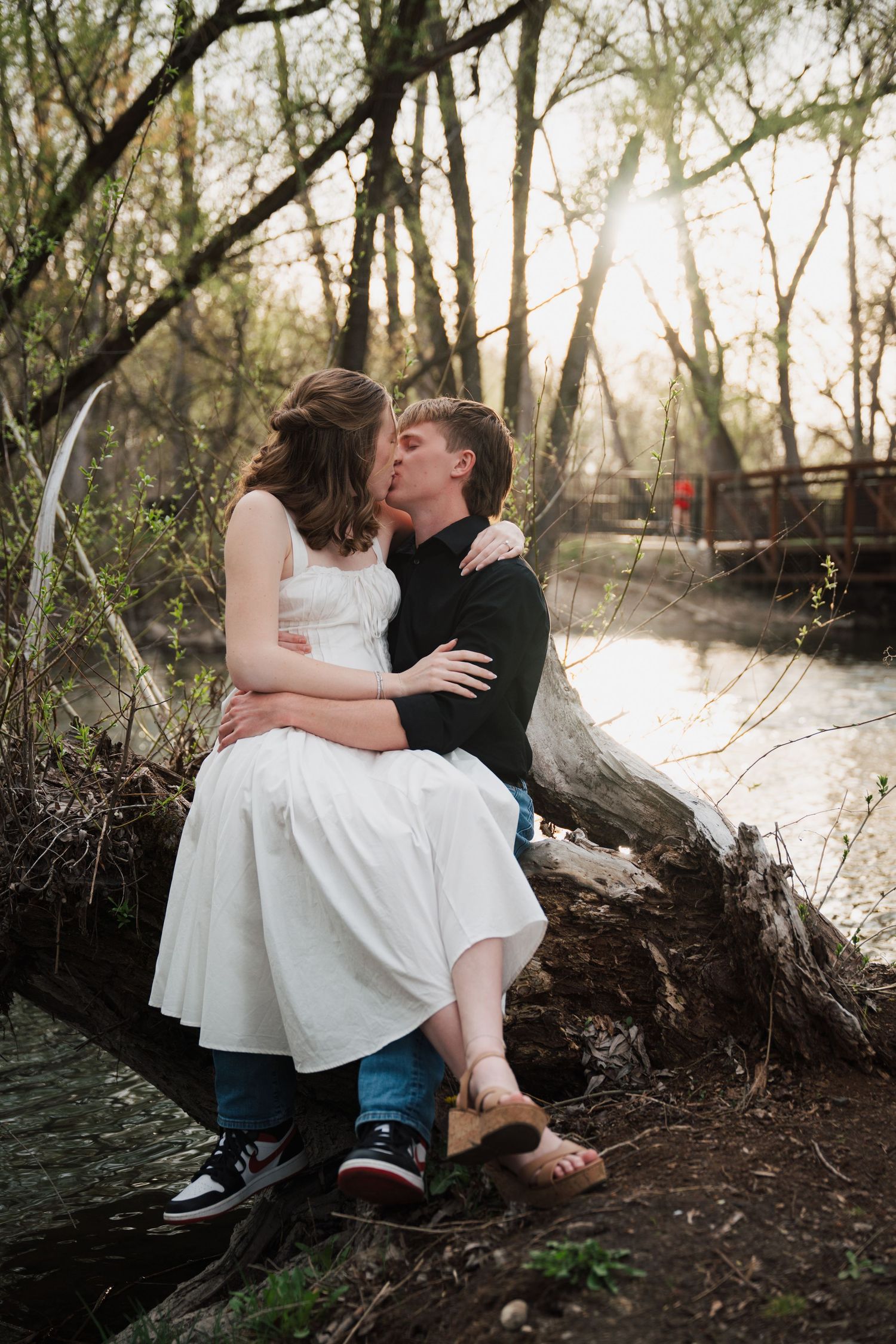 A couple shares an intimate moment while sitting on a fallen tree trunk by a river in a dreamy woodland setting.