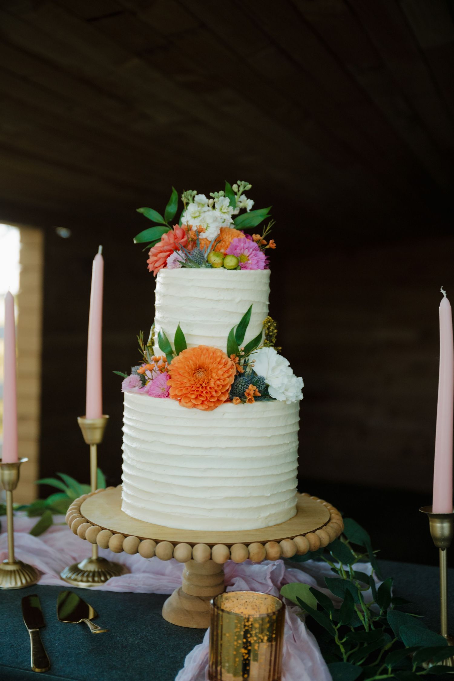 Lewis Lane Events in Nampa, Idaho. Two-tiered white wedding cake decorated with colorful flowers on a wooden stand with lit candles.
