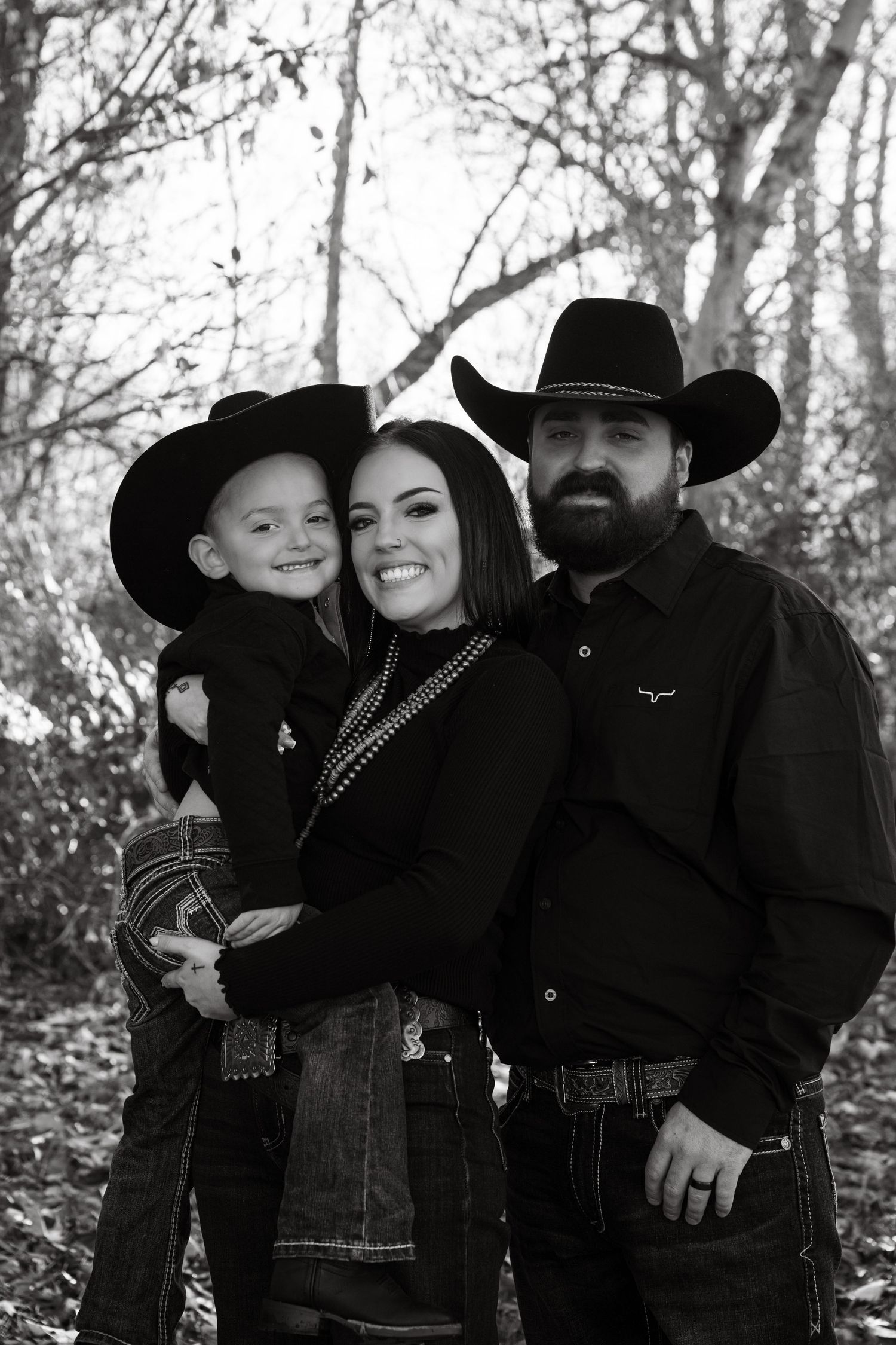 Black and white portrait of a family in western attire with cowboy hats standing together outdoors among trees.