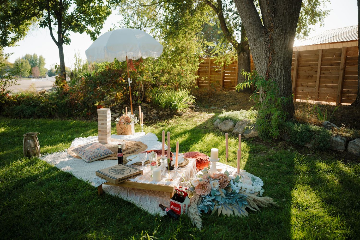 A romantic picnic setup in a backyard with blankets and decorative items on the grass.