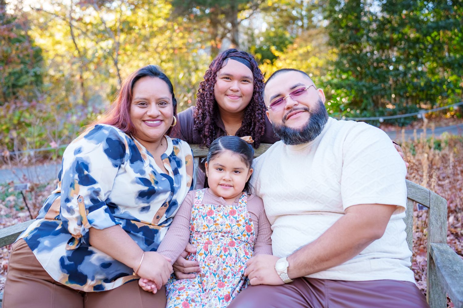 A family sits together outdoors during autumn with colorful fall foliage in the background.