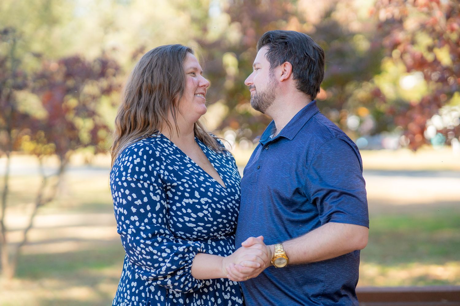 A couple shares a tender moment outdoors wearing blue clothing against an autumn backdrop with soft bokeh lighting.