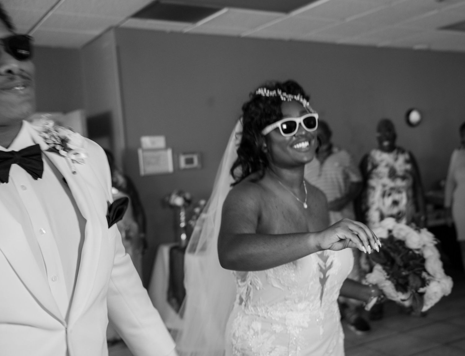 A joyful couple dancing at a wedding reception, with the bride wearing a white dress and sunglasses.