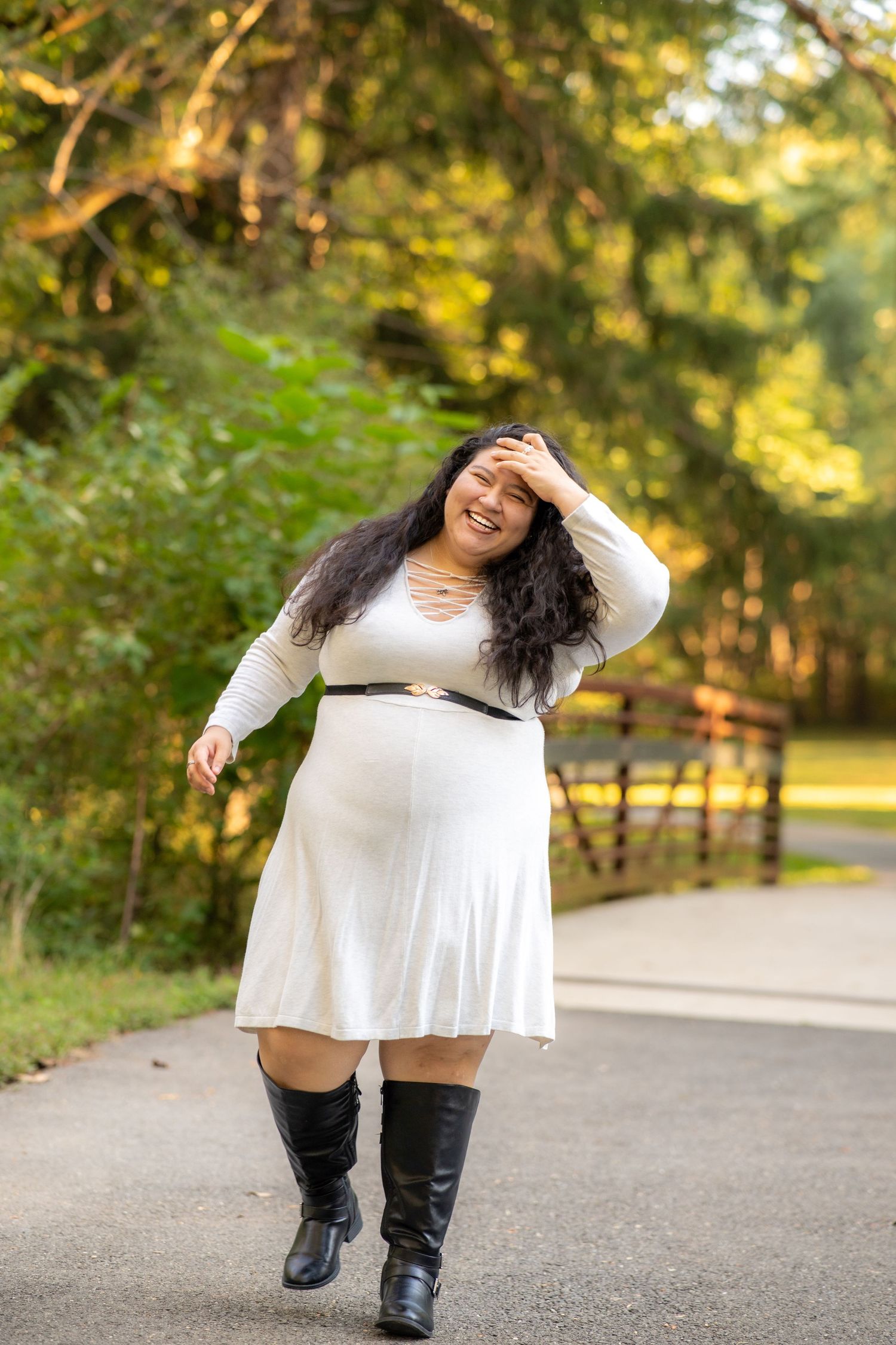 Someone wearing a white dress and tall boots dances expressively on a tree-lined path during sunset.