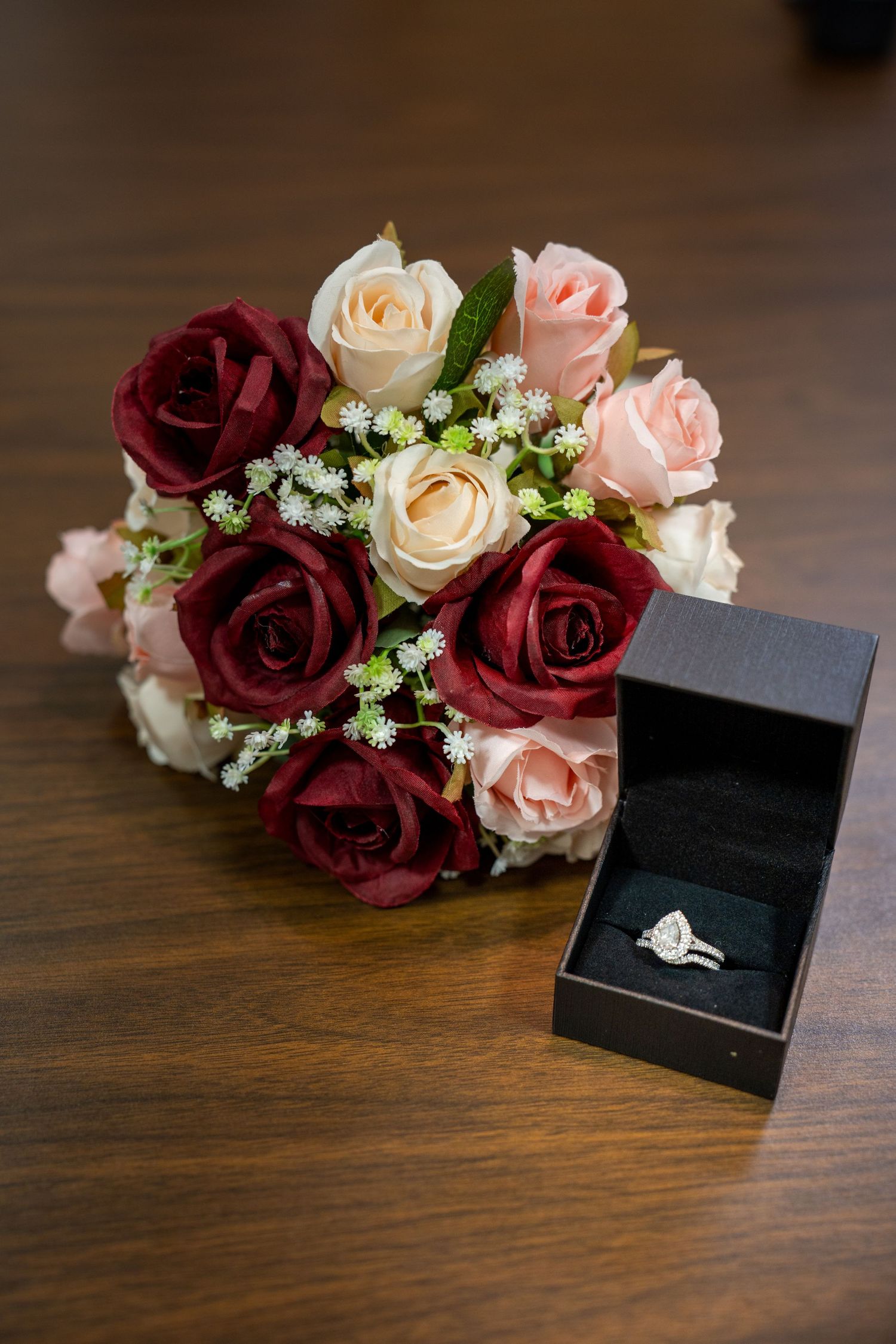 Engagement ring in black box with burgundy and pink roses arranged on wooden surface.
