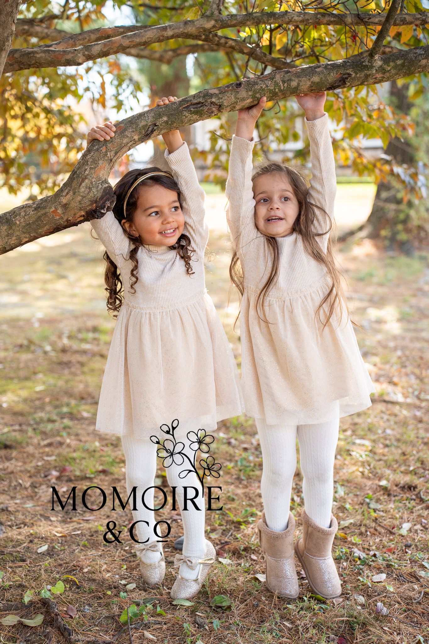 Two children in matching white dresses playing under a tree branch with autumn leaves.