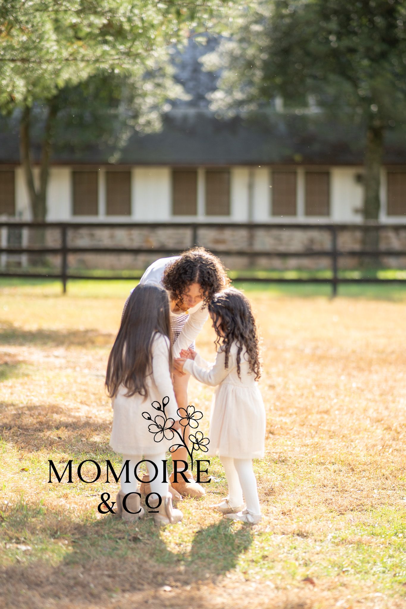 Two children in white dresses play together in a sunlit autumn park with fallen leaves on the ground.