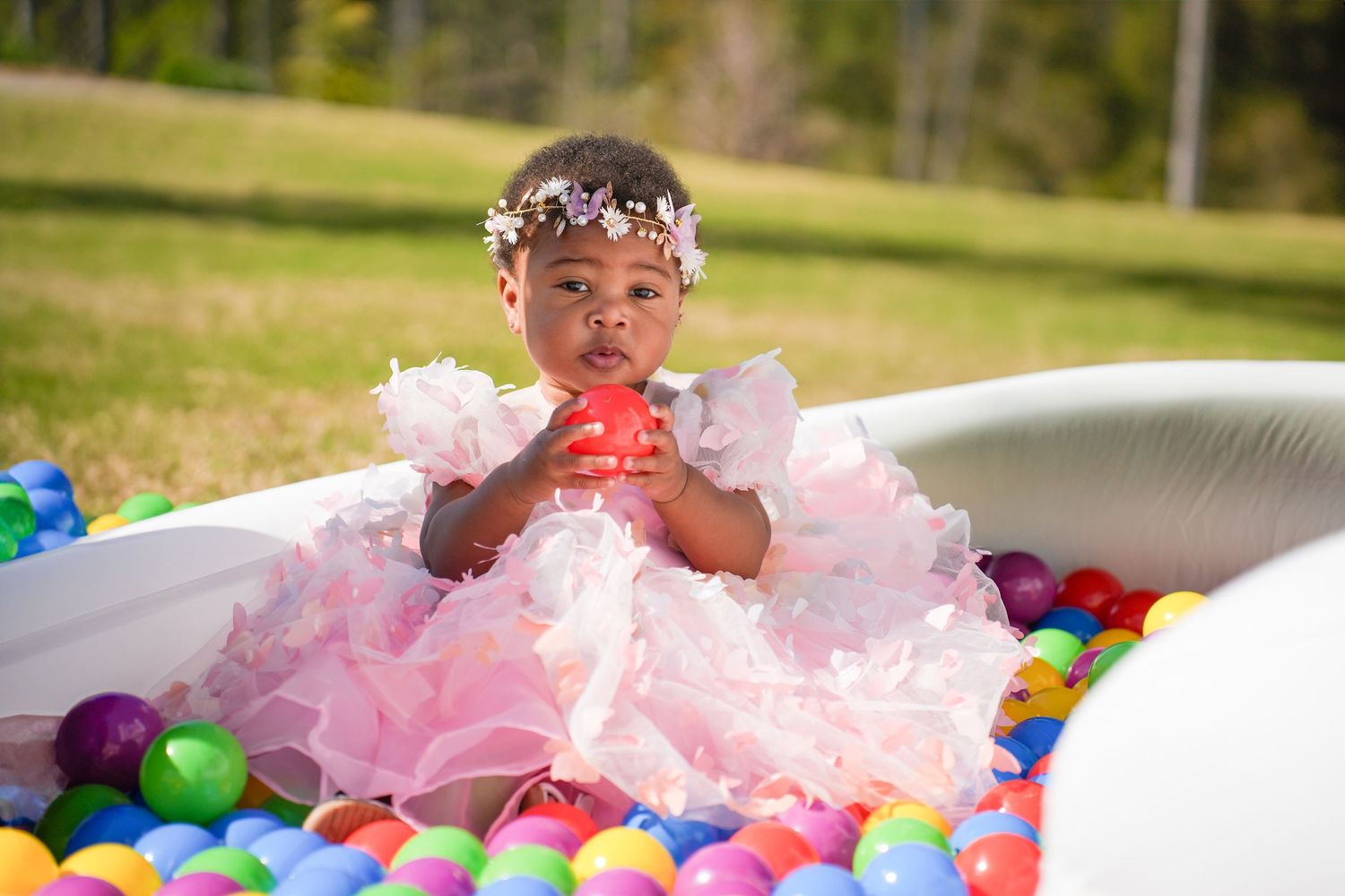 A child plays in a kiddie pool filled with colorful plastic balls and pink tulle decorations on a sunny day outdoors.