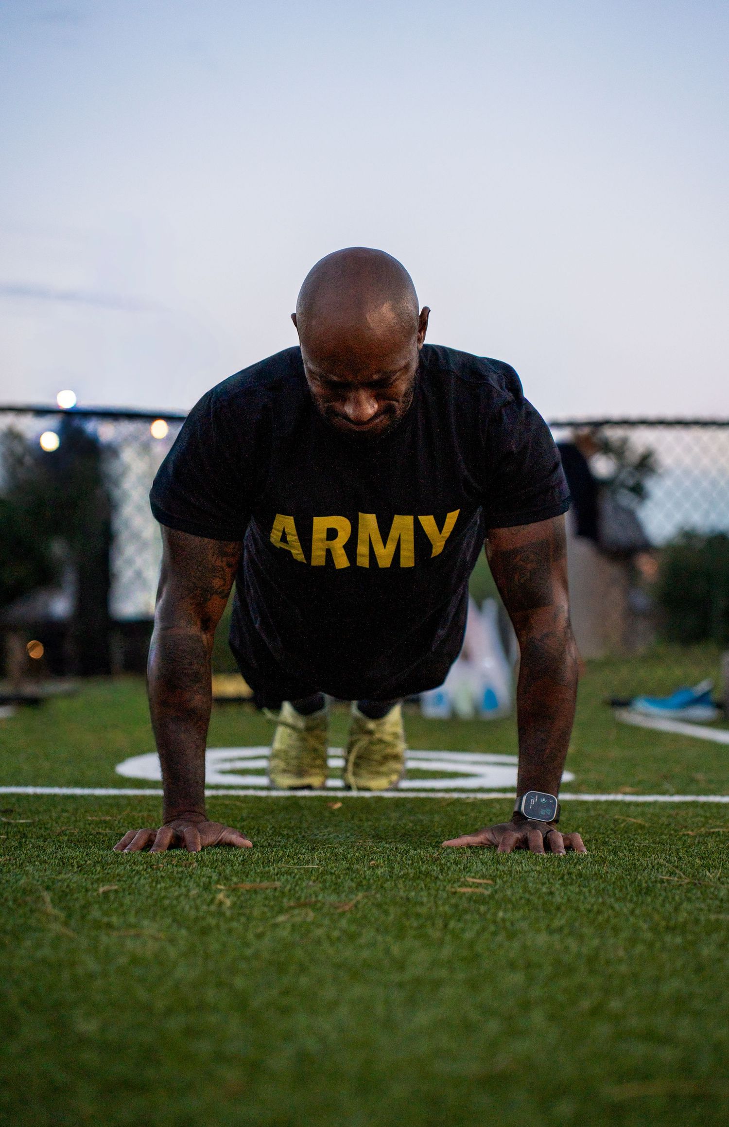 Person in Army t-shirt doing push-ups on grass field during outdoor training session at dusk.