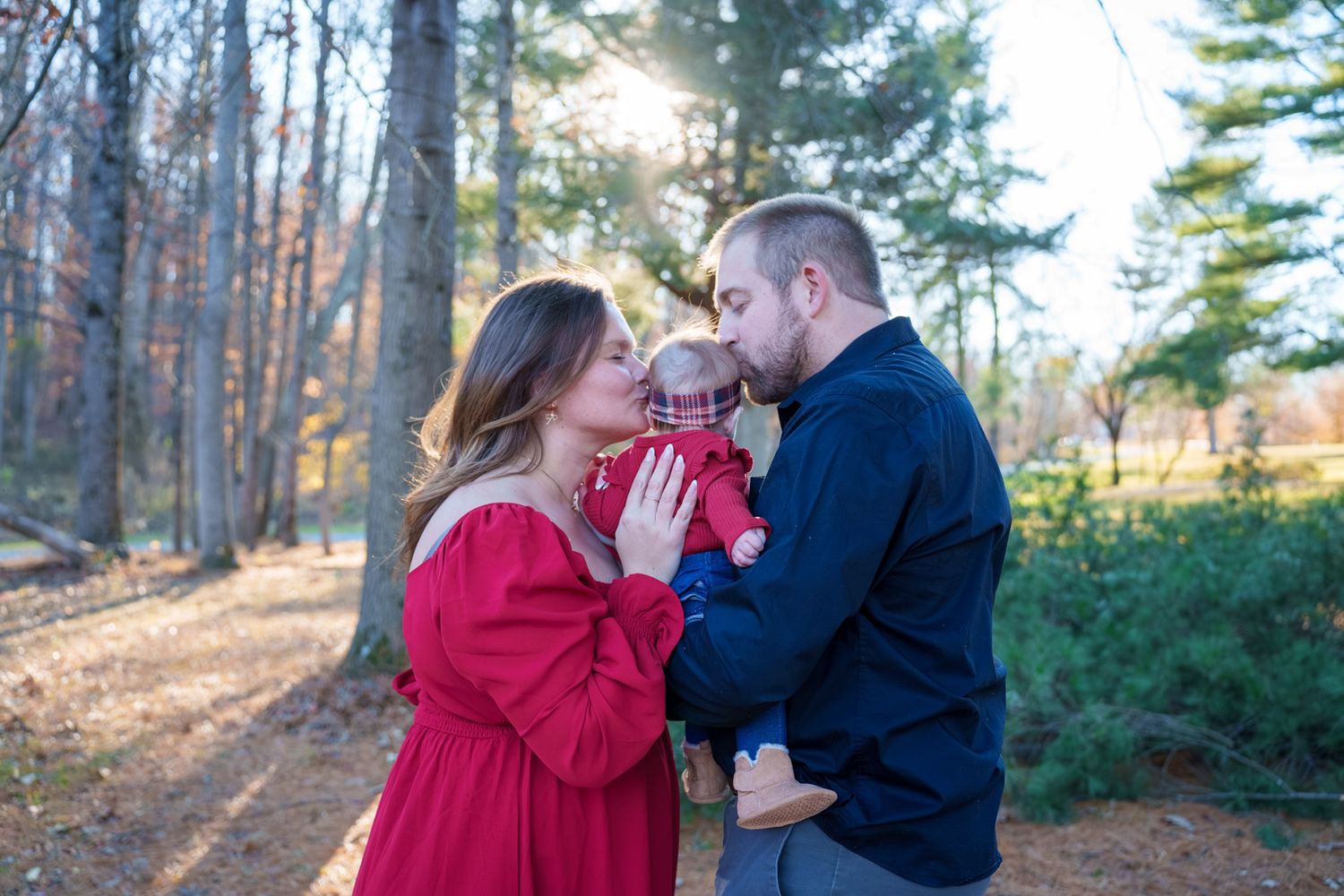 A couple in red and black attire shares romantic moments in a sunlit forest setting during an outdoor photoshoot.