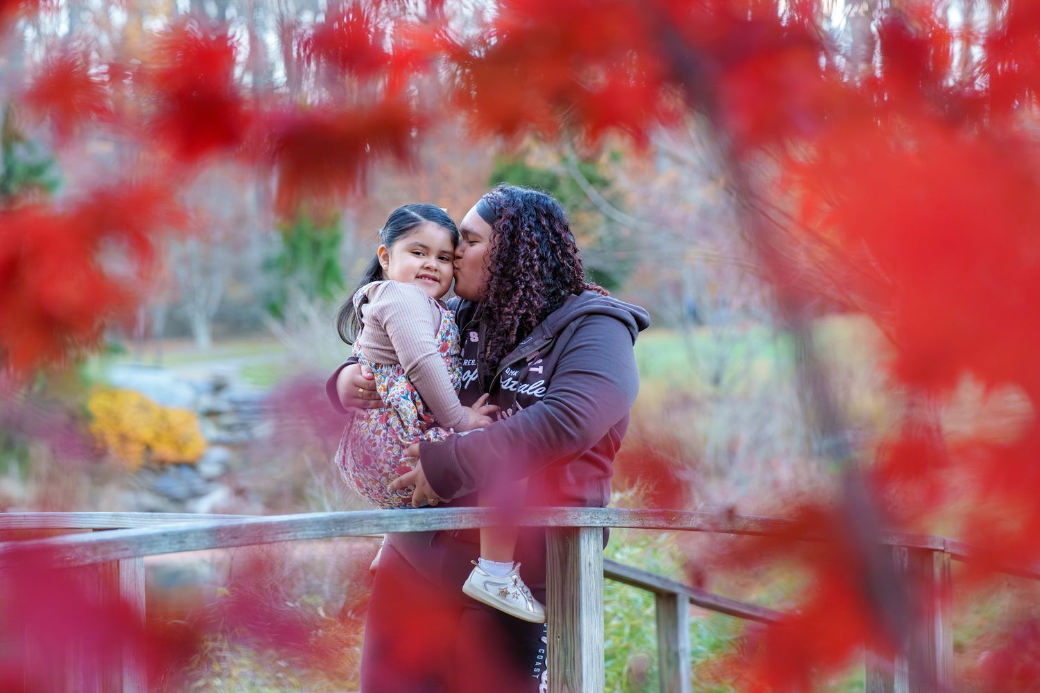 A couple shares a romantic moment surrounded by vibrant red autumn foliage in a park setting.