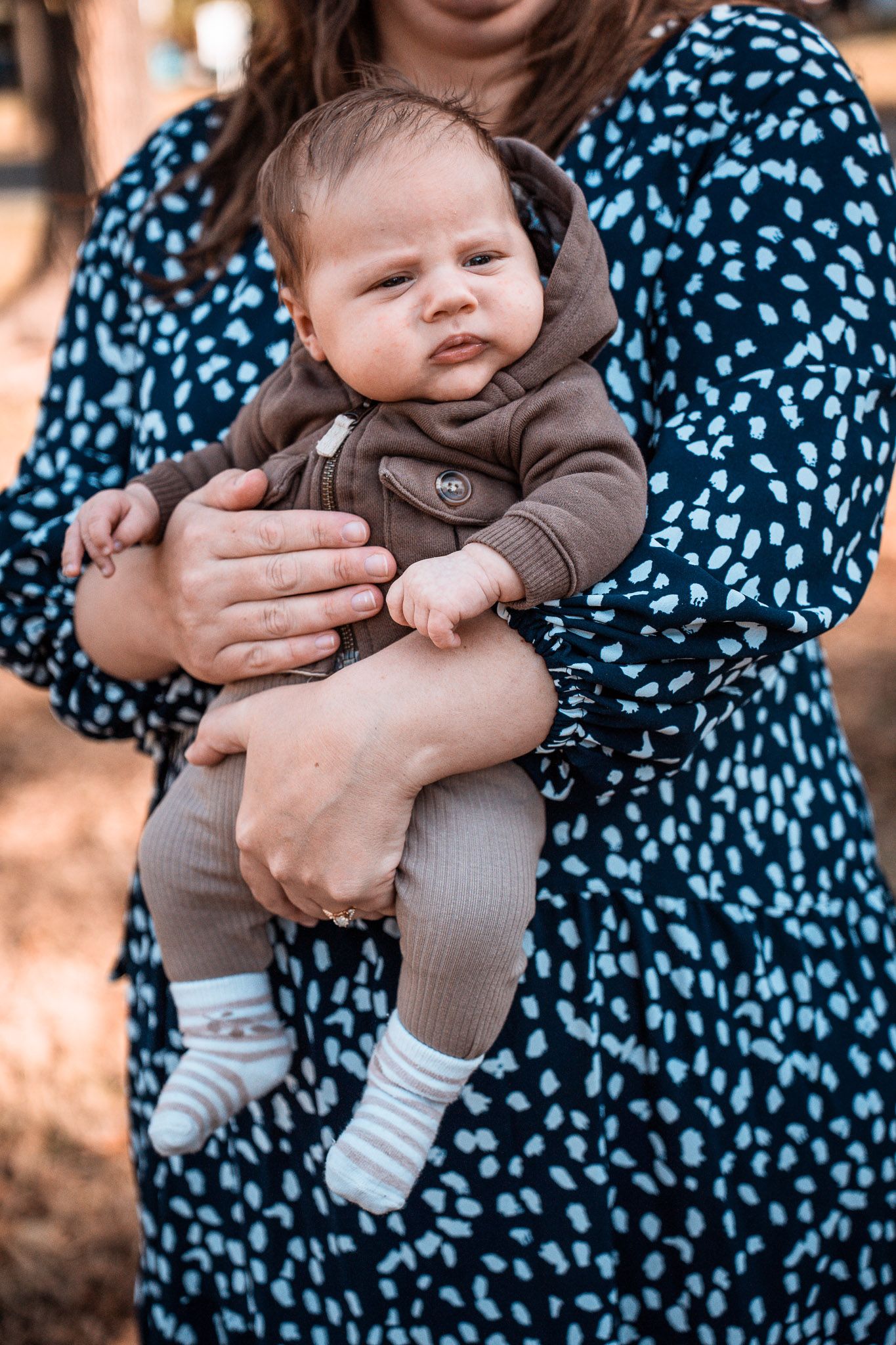 A woman in a blue patterned dress holds a small baby wearing a brown outfit against a blurred background.