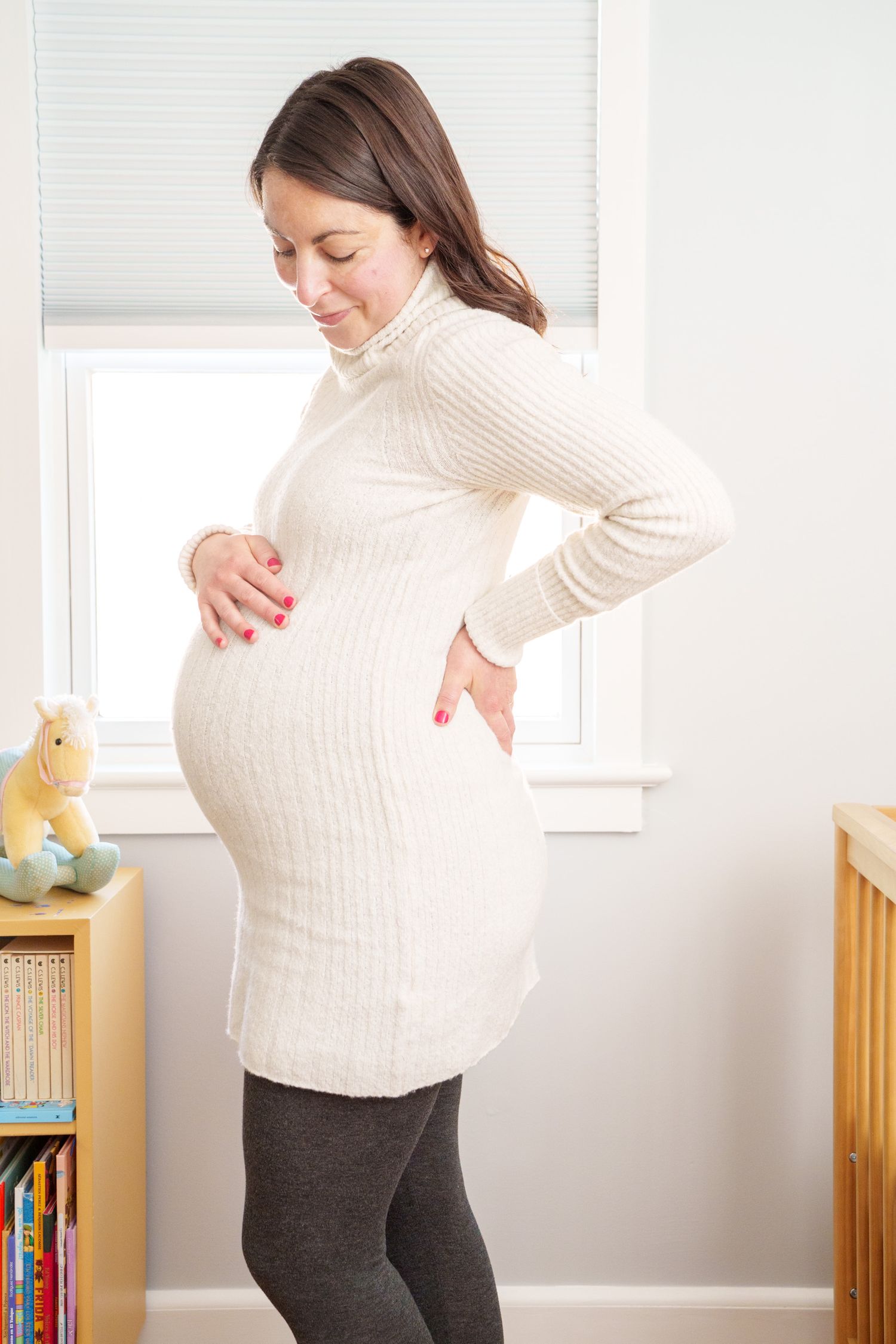 Pregnant woman in white sweater stands by window showing progression of growing baby bump in multiple poses.