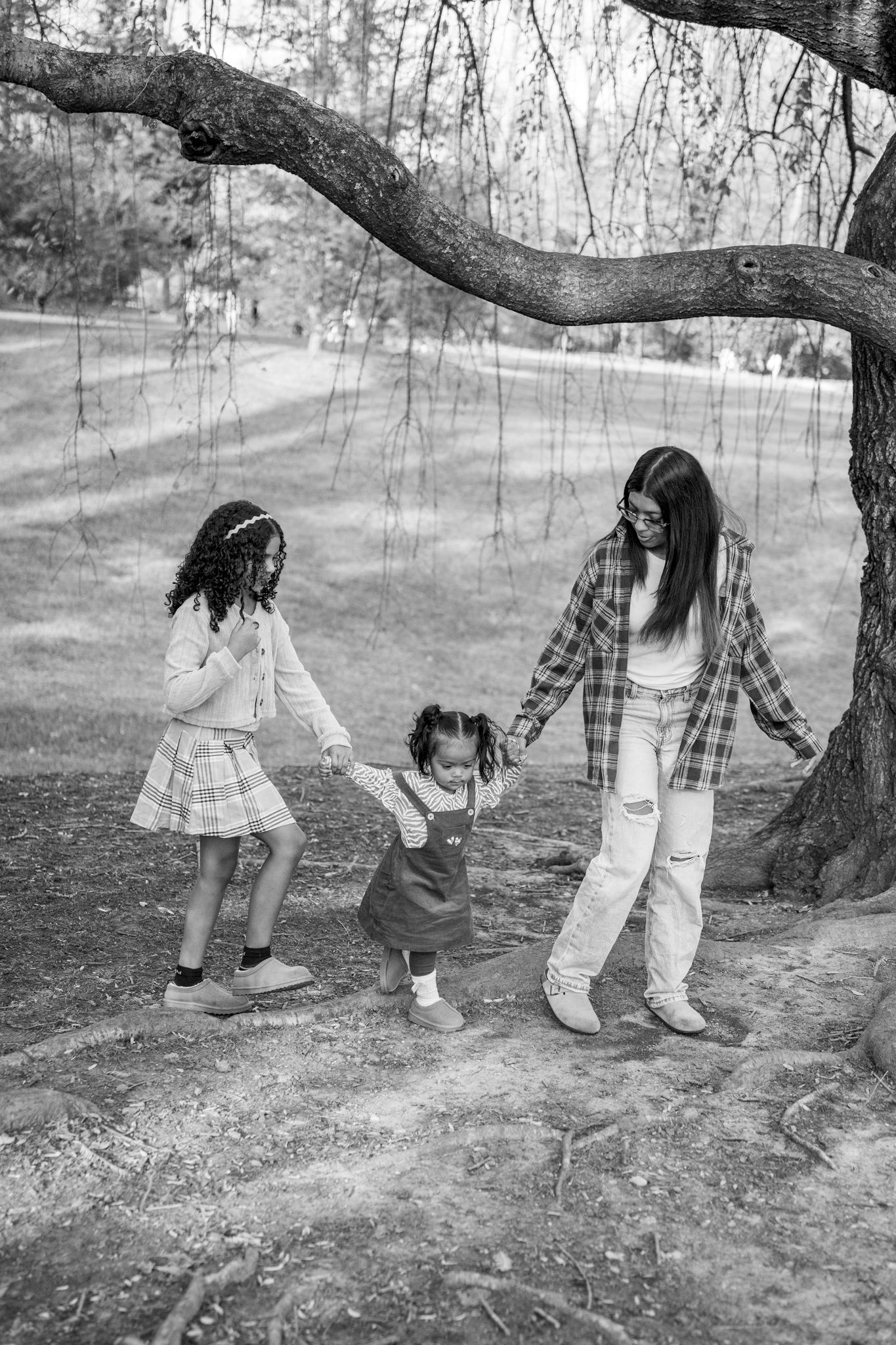 Black and white photo showing three people holding hands and dancing under a large tree branch in a park setting.
