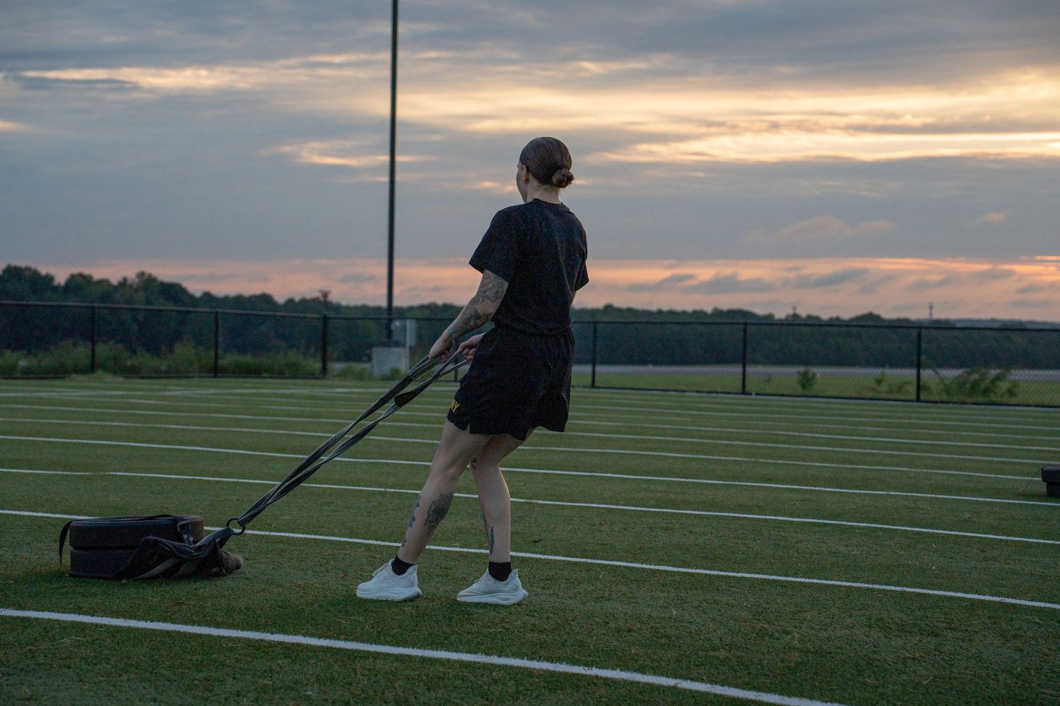 Person in black outfit pulling equipment on a sports field at sunset with dramatic sky in background.