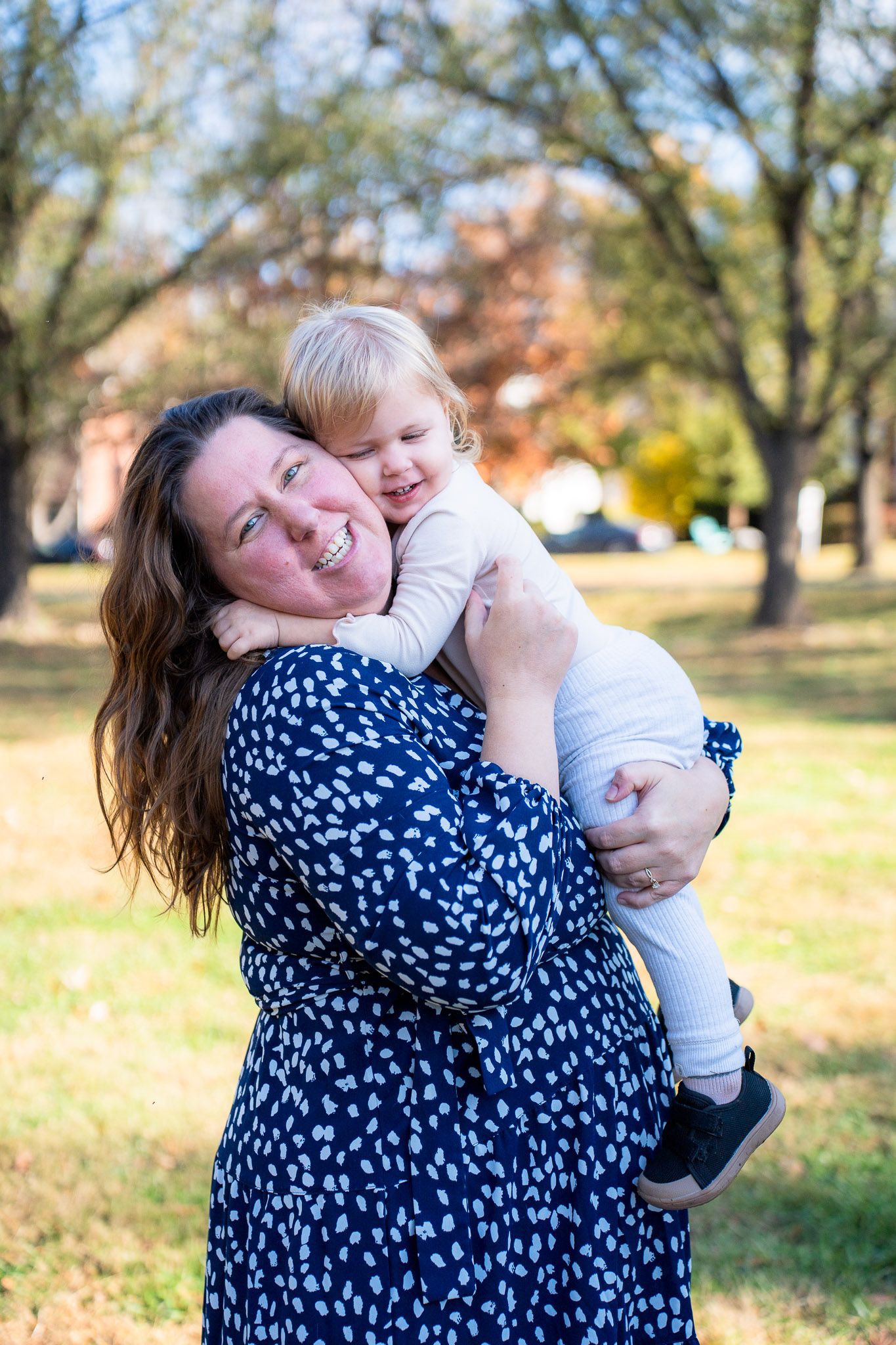 A woman in a navy floral dress plays and laughs while holding a baby outdoors in autumn.