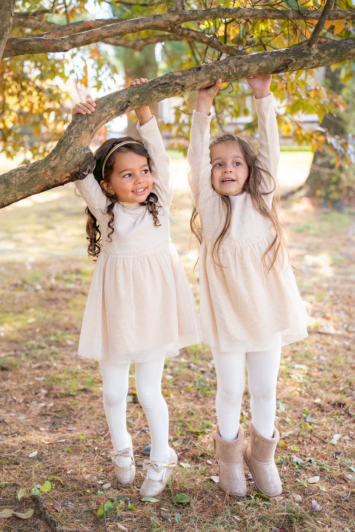 Two children in matching white dresses play beneath autumn tree branches on fallen leaves.