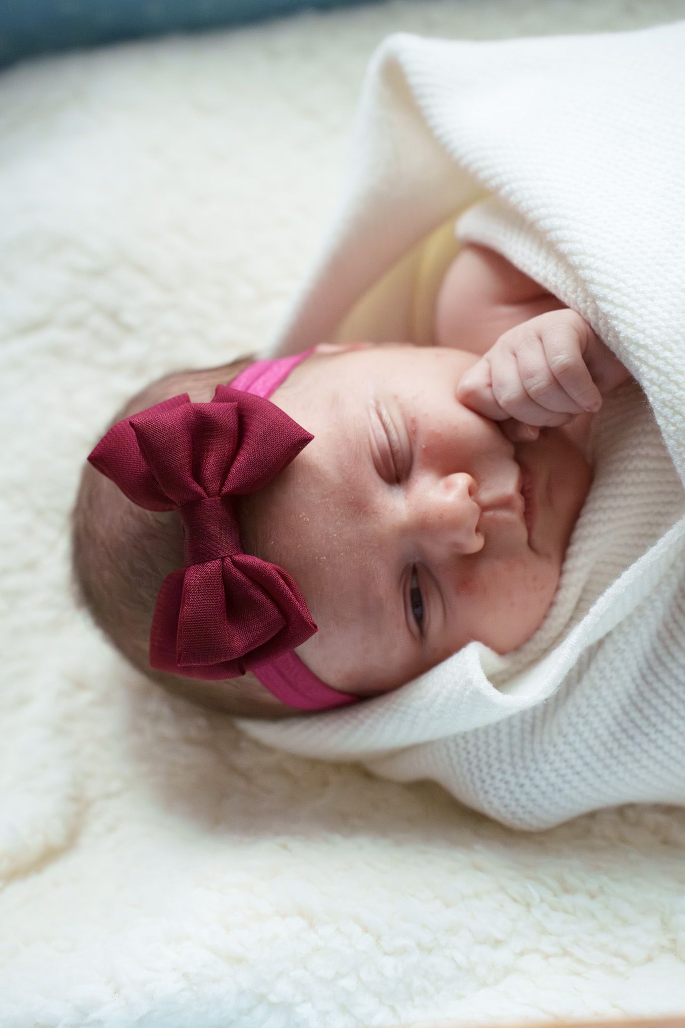 A sleeping newborn baby wearing a burgundy bow headband and white knit sweater lies on soft cream bedding.