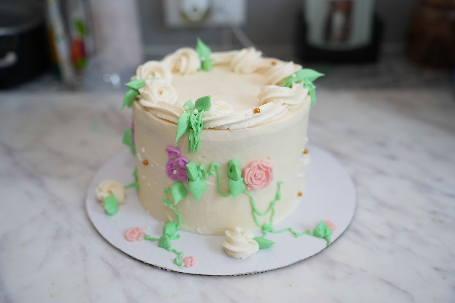A white frosted cake decorated with delicate pink sugar roses and green leaves on a marble countertop.