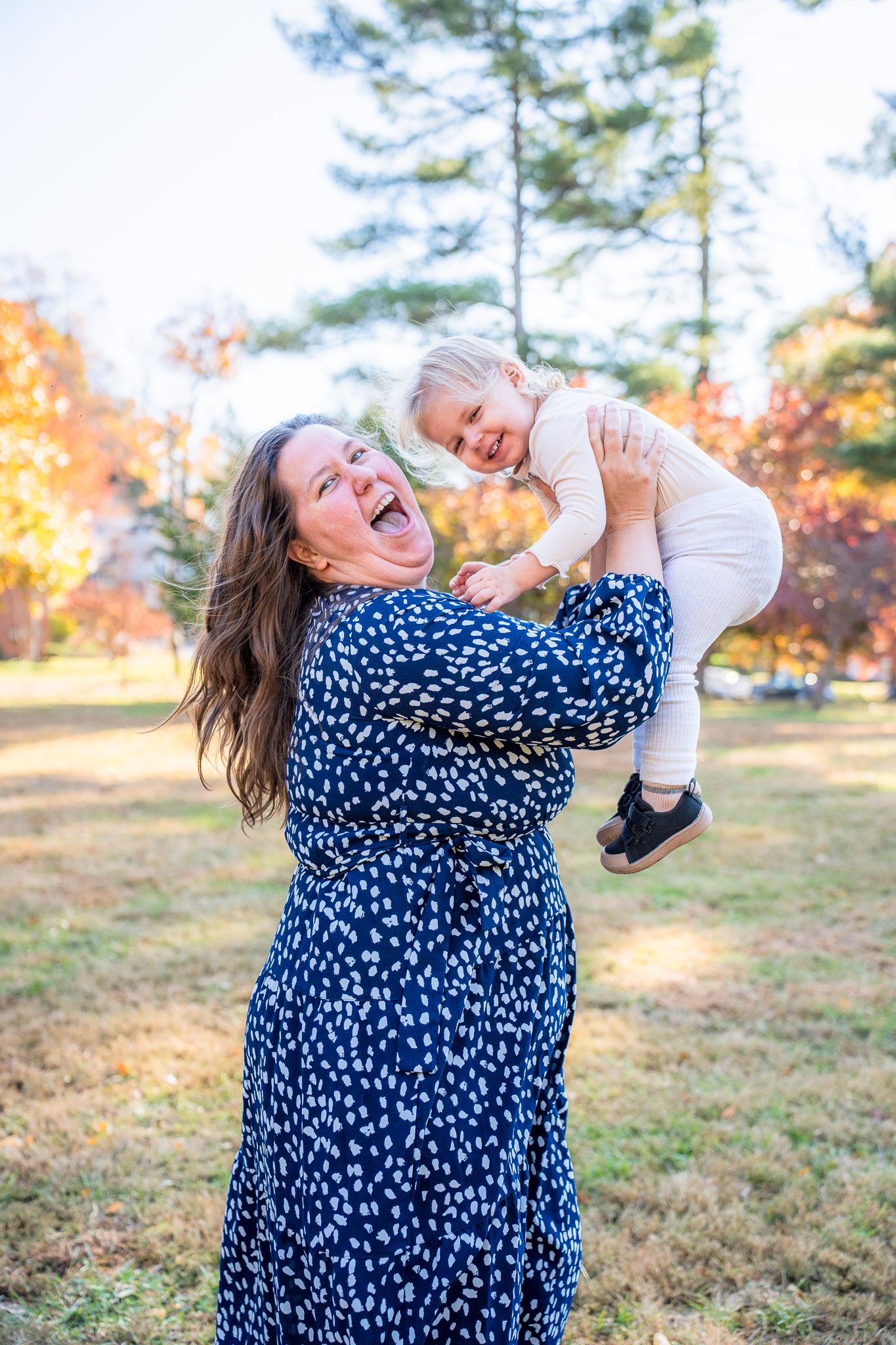 A person in a blue polka dot dress dances joyfully in a sunny outdoor field with autumn trees in the background.