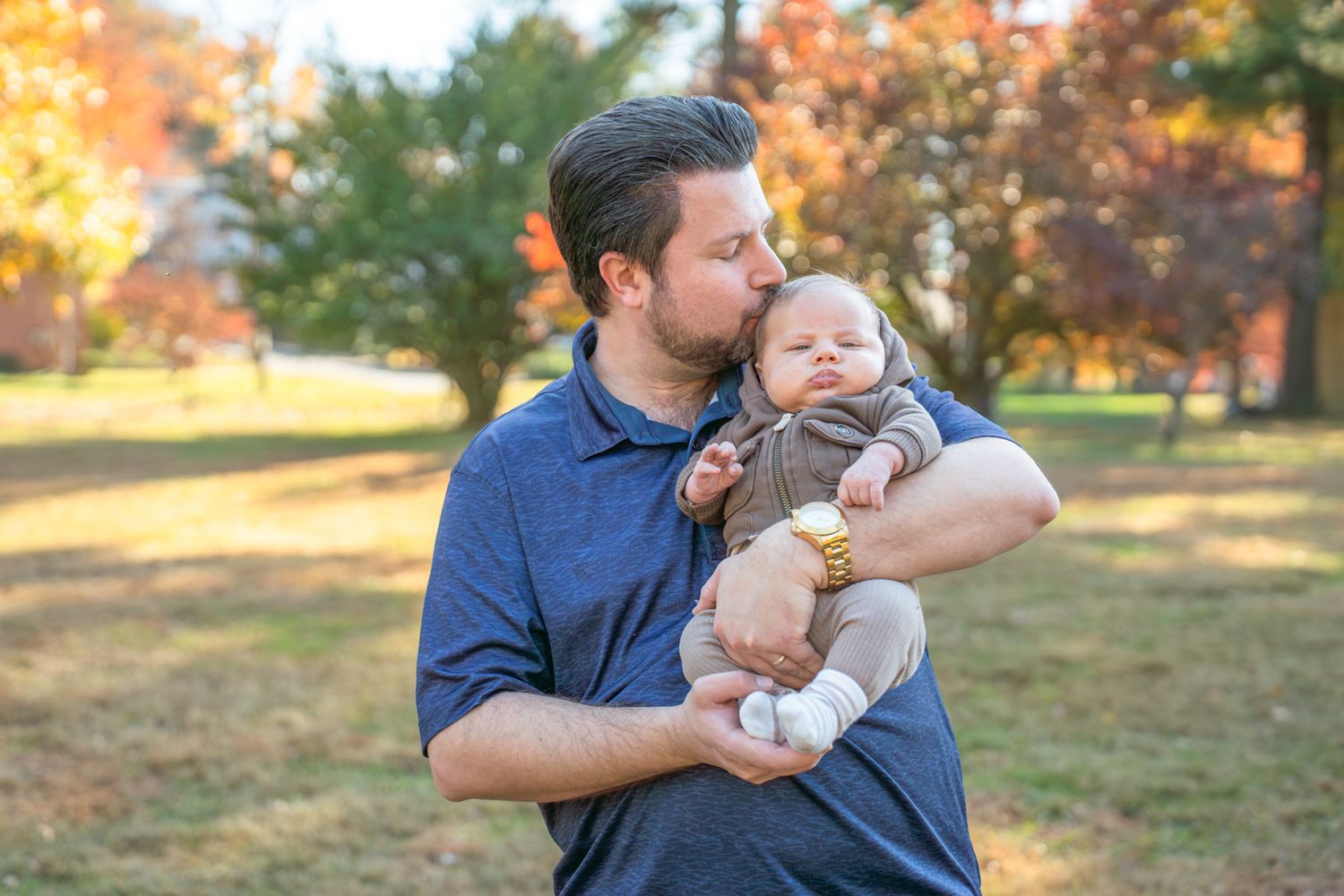 Parent and child share tender moments outdoors during autumn with golden sunlight and fall foliage in background.