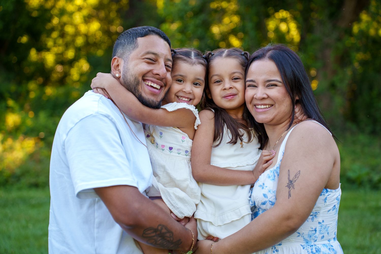 Happy family of four smiling outdoors, parents holding two young girls in white dresses against a green park background.