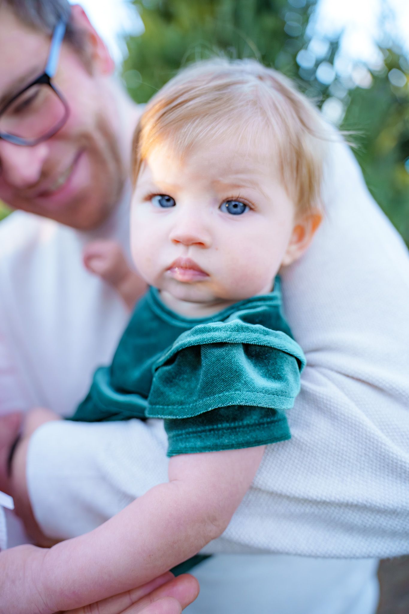 A blue-eyed baby in a green outfit being held by an adult wearing glasses outdoors.