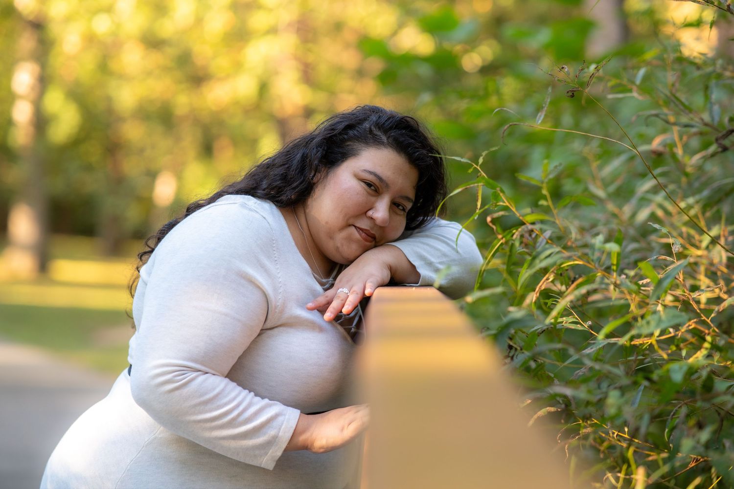 A person in a white sweater poses thoughtfully in a sunlit park setting with autumn foliage in the background.