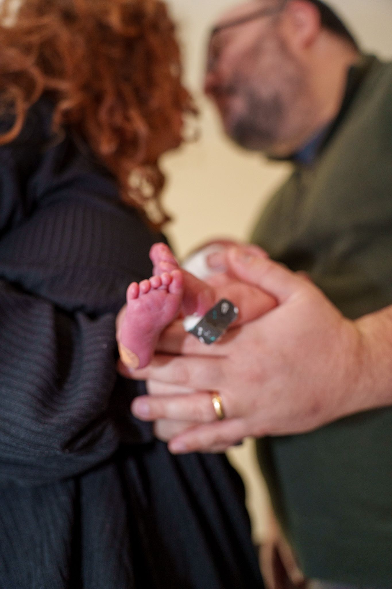 Close-up of tiny pink baby foot being gently held by adult hands wearing a gold wedding ring.