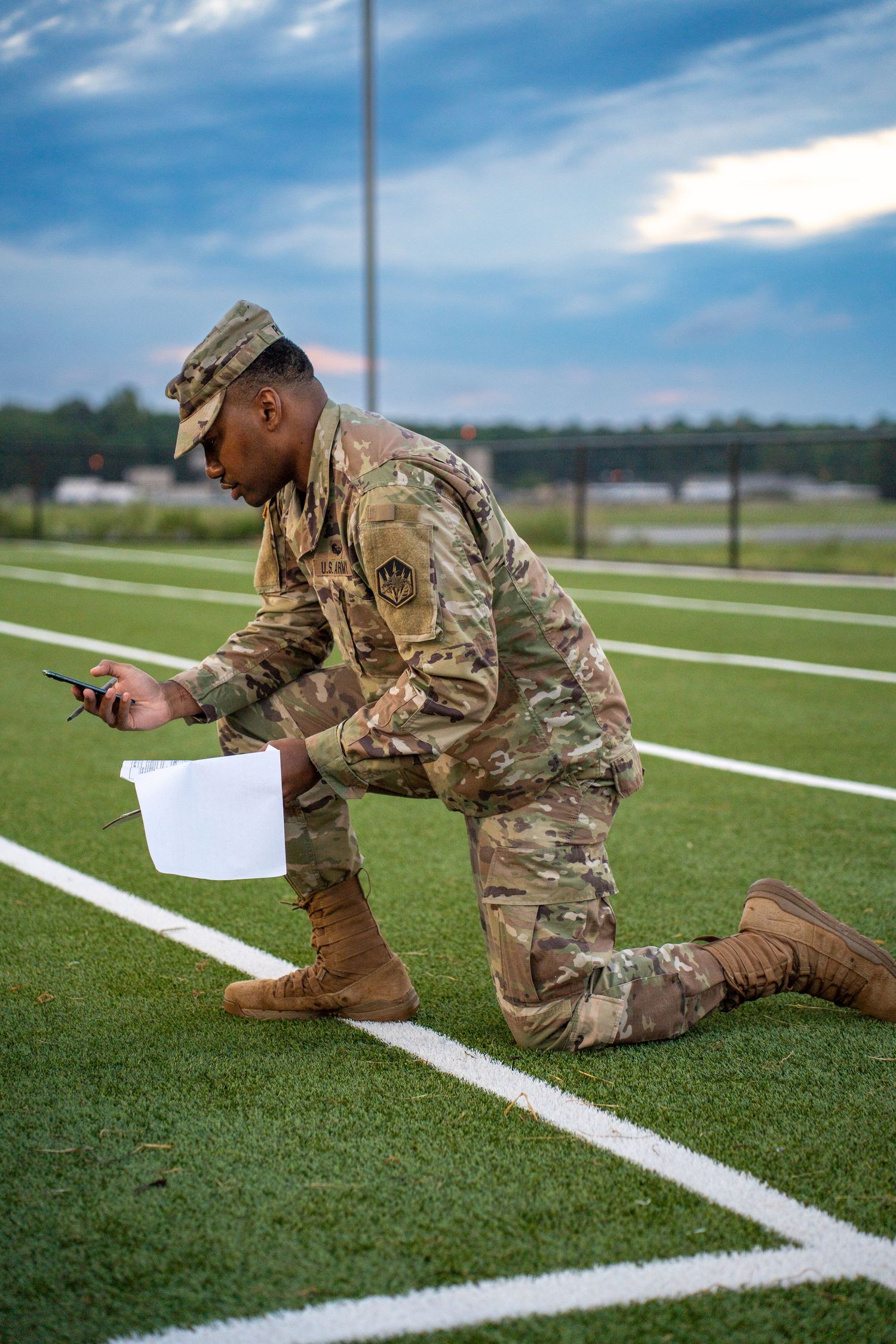 Military service member in camouflage uniform measures lines on athletic field with measuring tape.