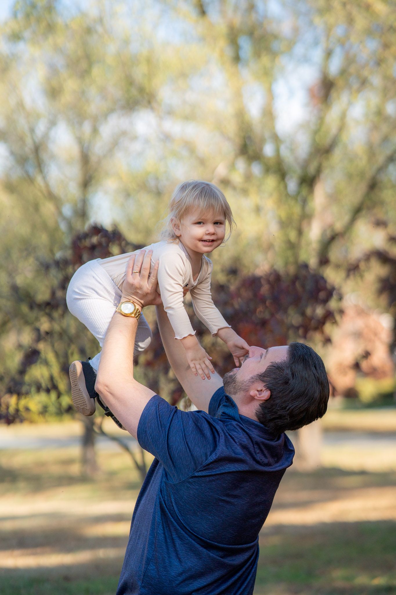 A parent lifts a small child joyfully into the air in an outdoor park setting with trees in the background.