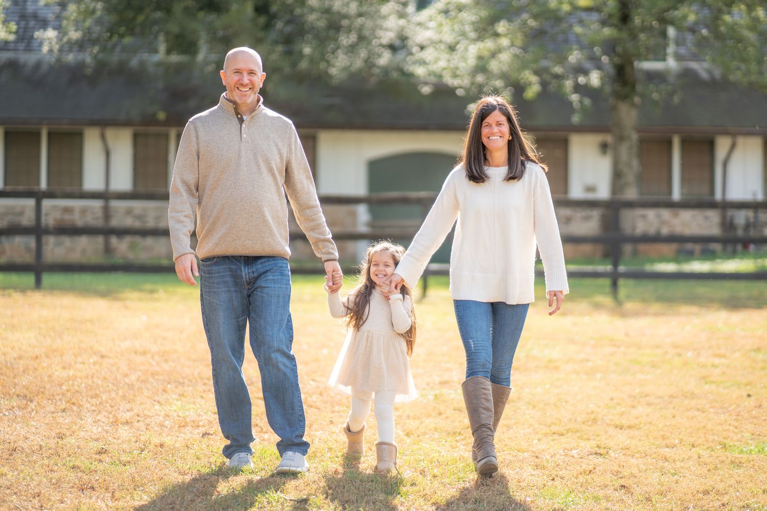 Family walks together holding hands on autumn grass in front of rustic barn on sunny afternoon.