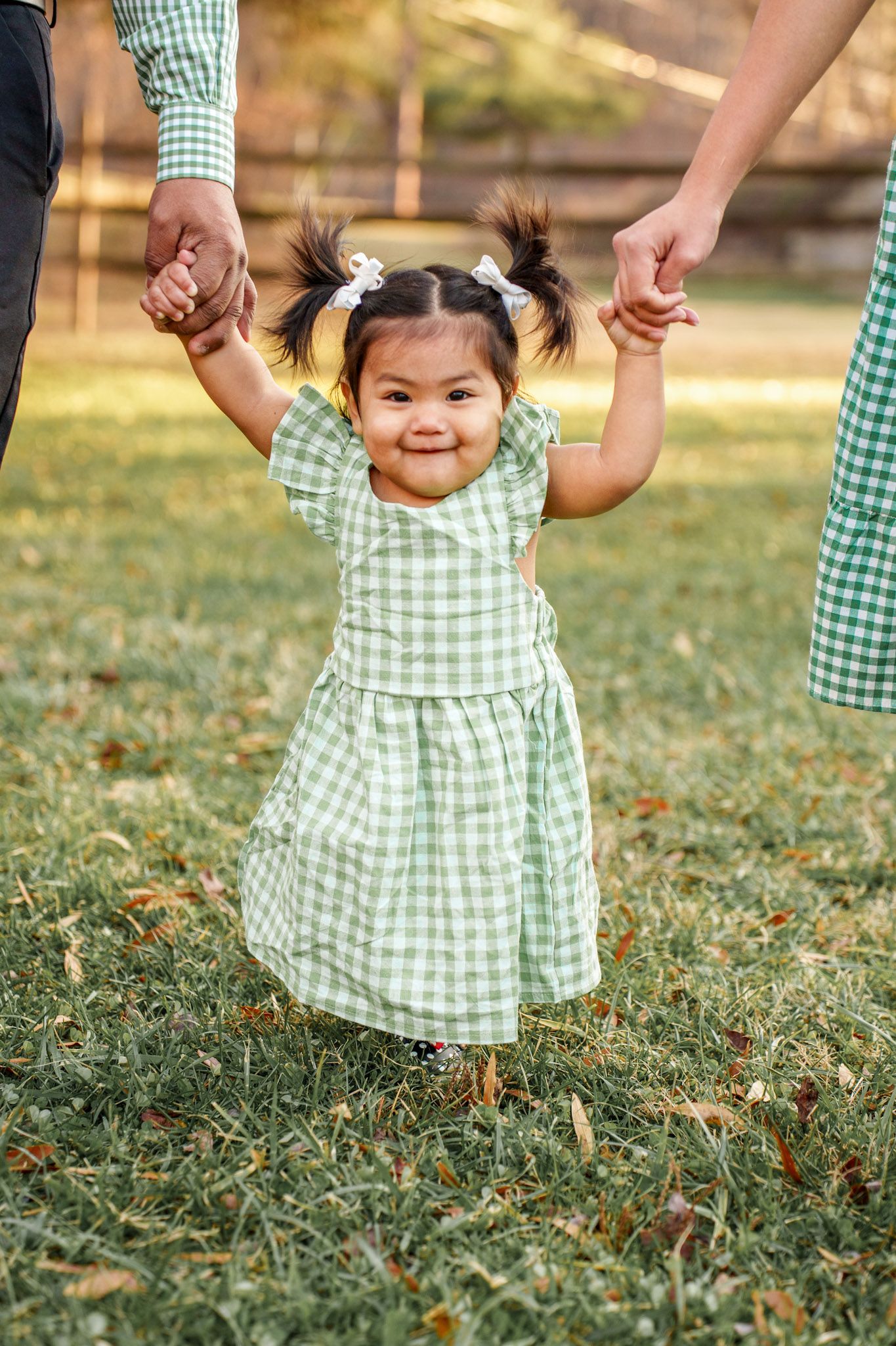 A child in a green gingham dress holds hands and walks through grass during a sunny outdoor photo session.
