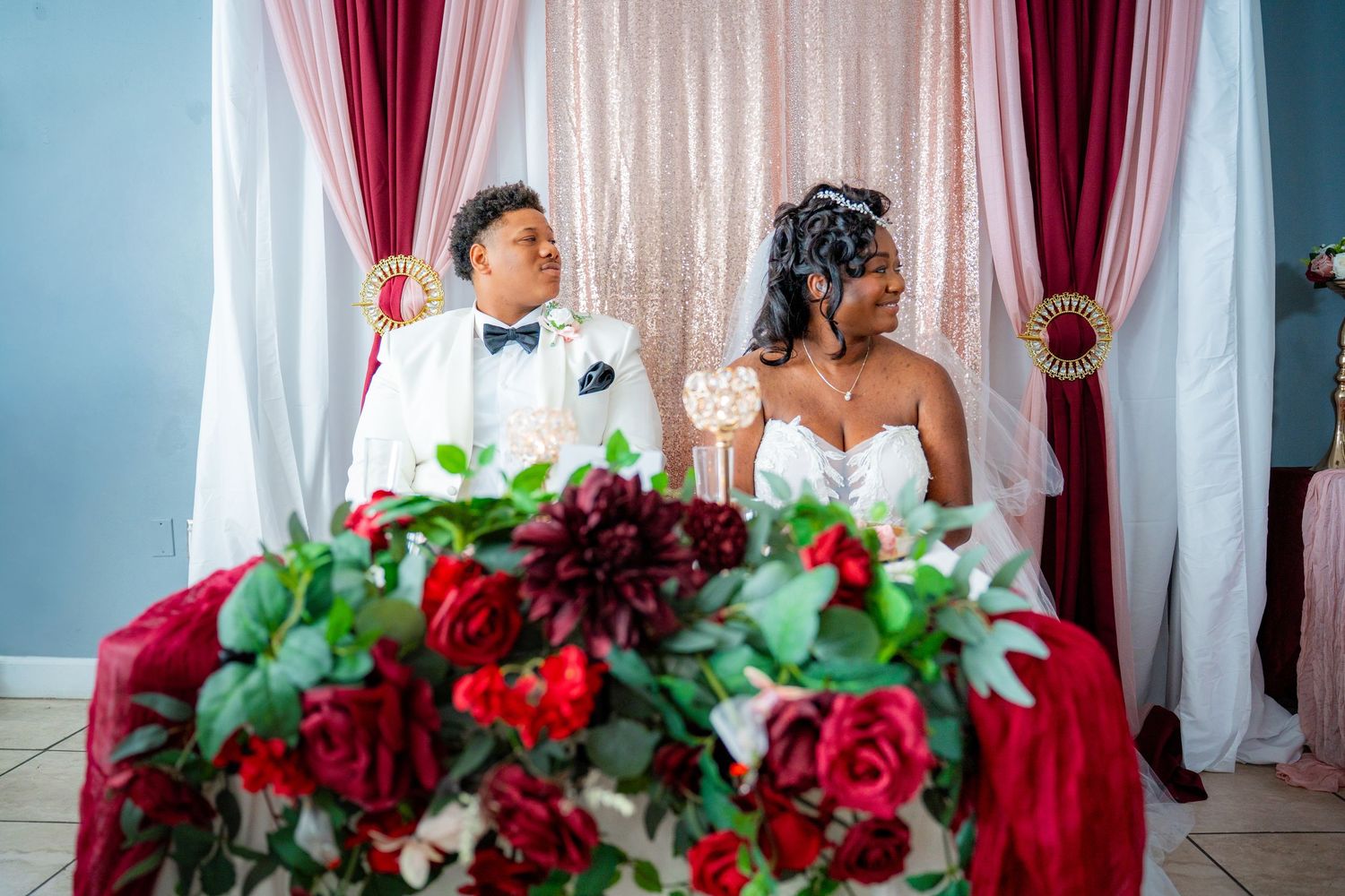 Wedding reception venue decorated with red and white floral arrangements and burgundy drapes.